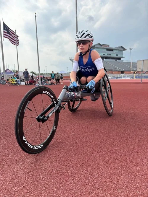 Boy waering a helmet, sunglasses and a blue shirt in a racing wheelchair on a track. 