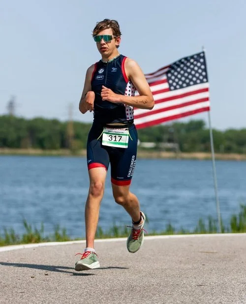 Man running a road race wearing a blue singlet and an American flag in the background.