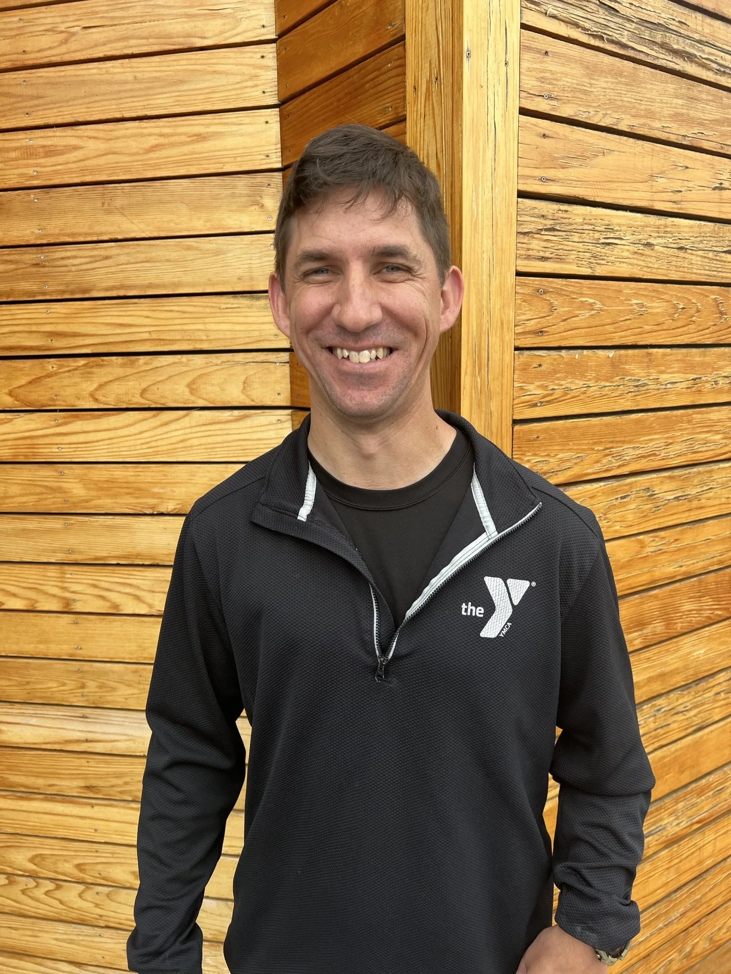 Man wearing a black athletic jacket with a white YMCA logo, posing outdoors against a background of warm-toned wooden panels