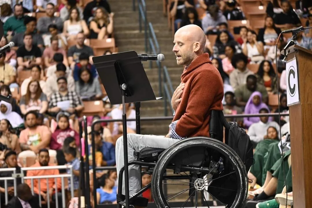 A man in a wheelchair is speaking or reading from a podium at a large indoor gathering or conference, with a diverse audience seated behind him.