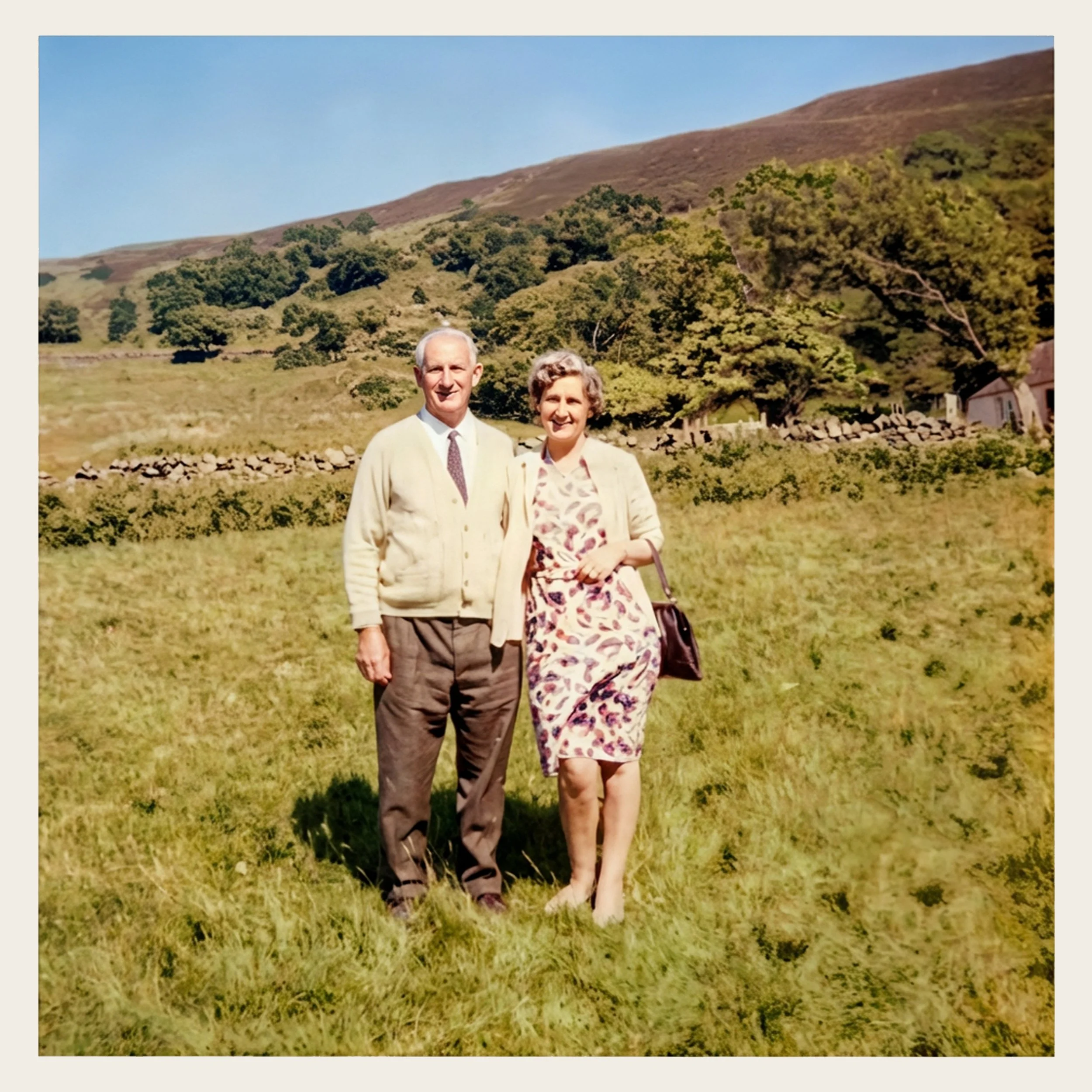 An elderly man and woman standing together in a grassy field with hills and trees in the background, on a sunny day.