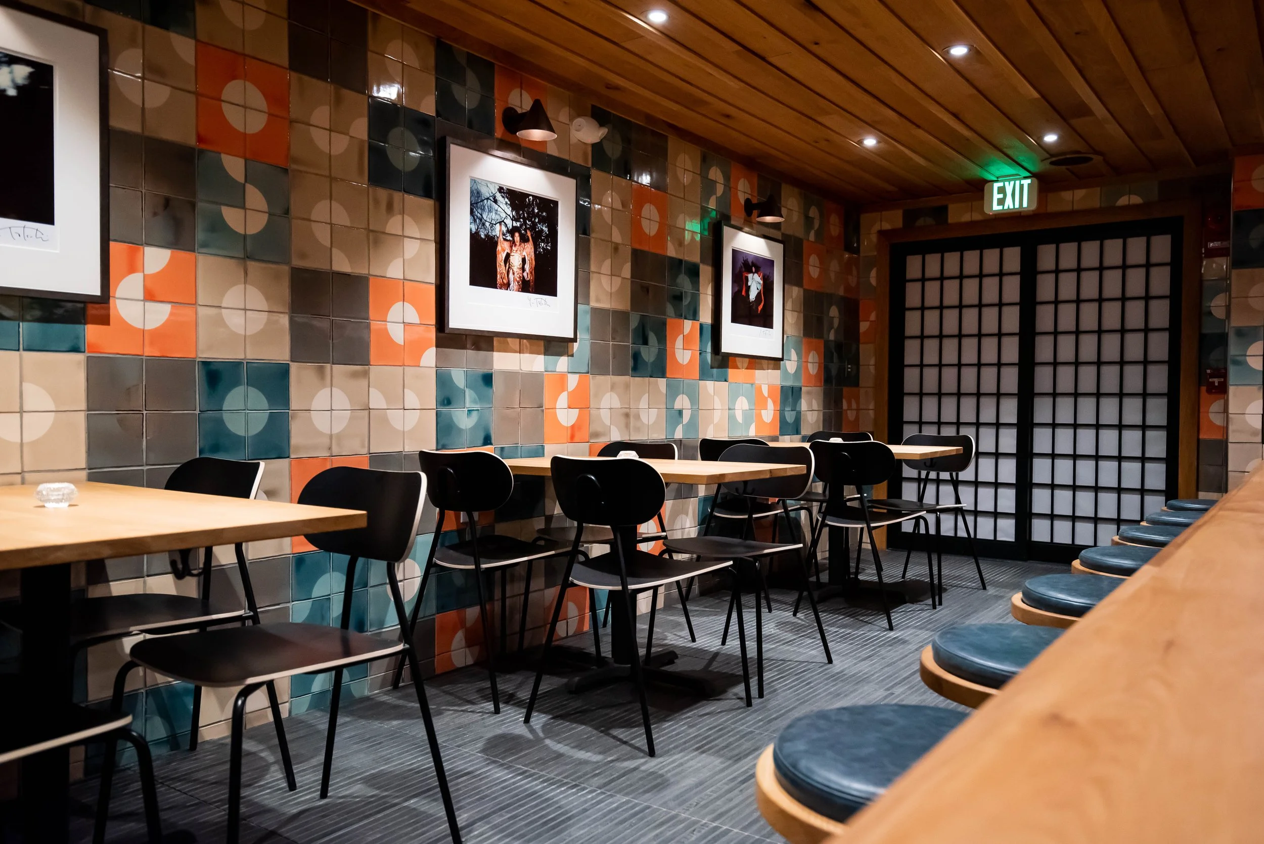 Sala interna del ristorante Kappo con pareti piastrellate geometriche, sedie nere, tavoli in legno chiaro e illuminazione soffusa.