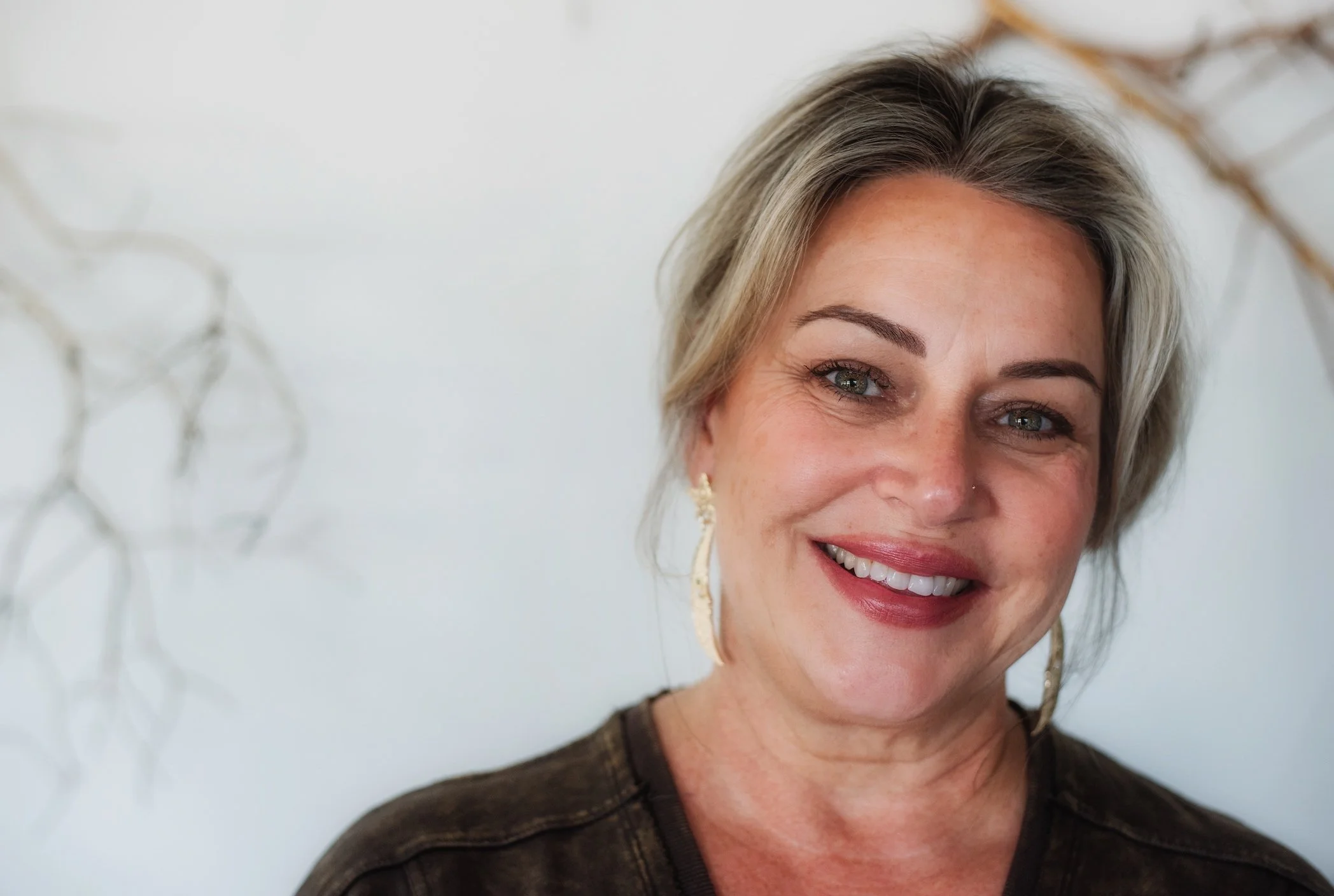 Close-up of a middle-aged woman with short gray hair, smiling, wearing earrings and a brown top, with a light background.