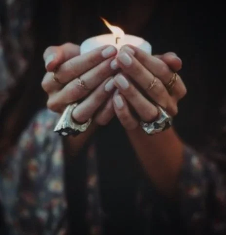 Person holding a lit white candle with both hands, wearing rings on fingers.