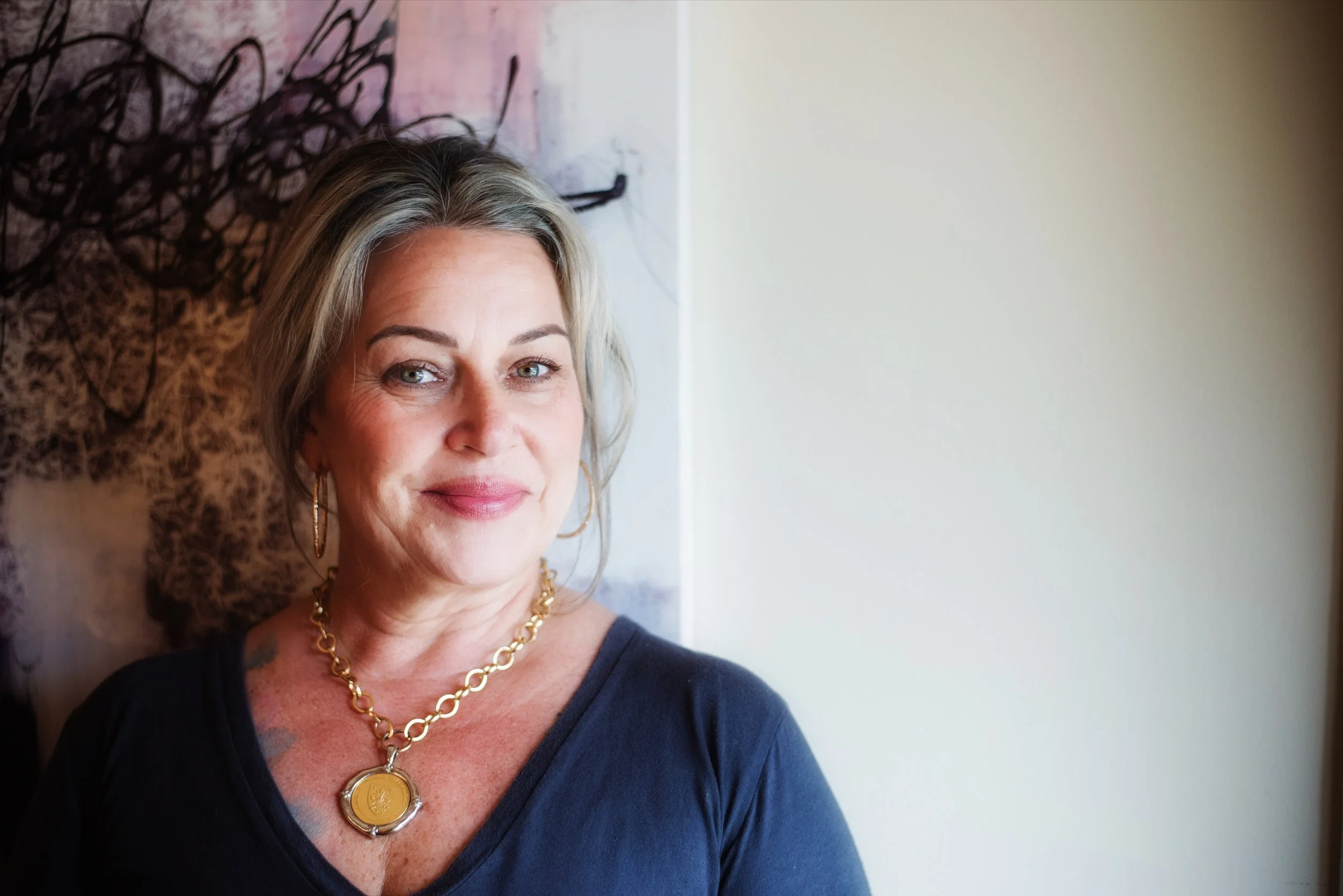 Portrait of a smiling middle-aged woman with gray hair, wearing gold jewelry, standing near an abstract artwork on a wall.