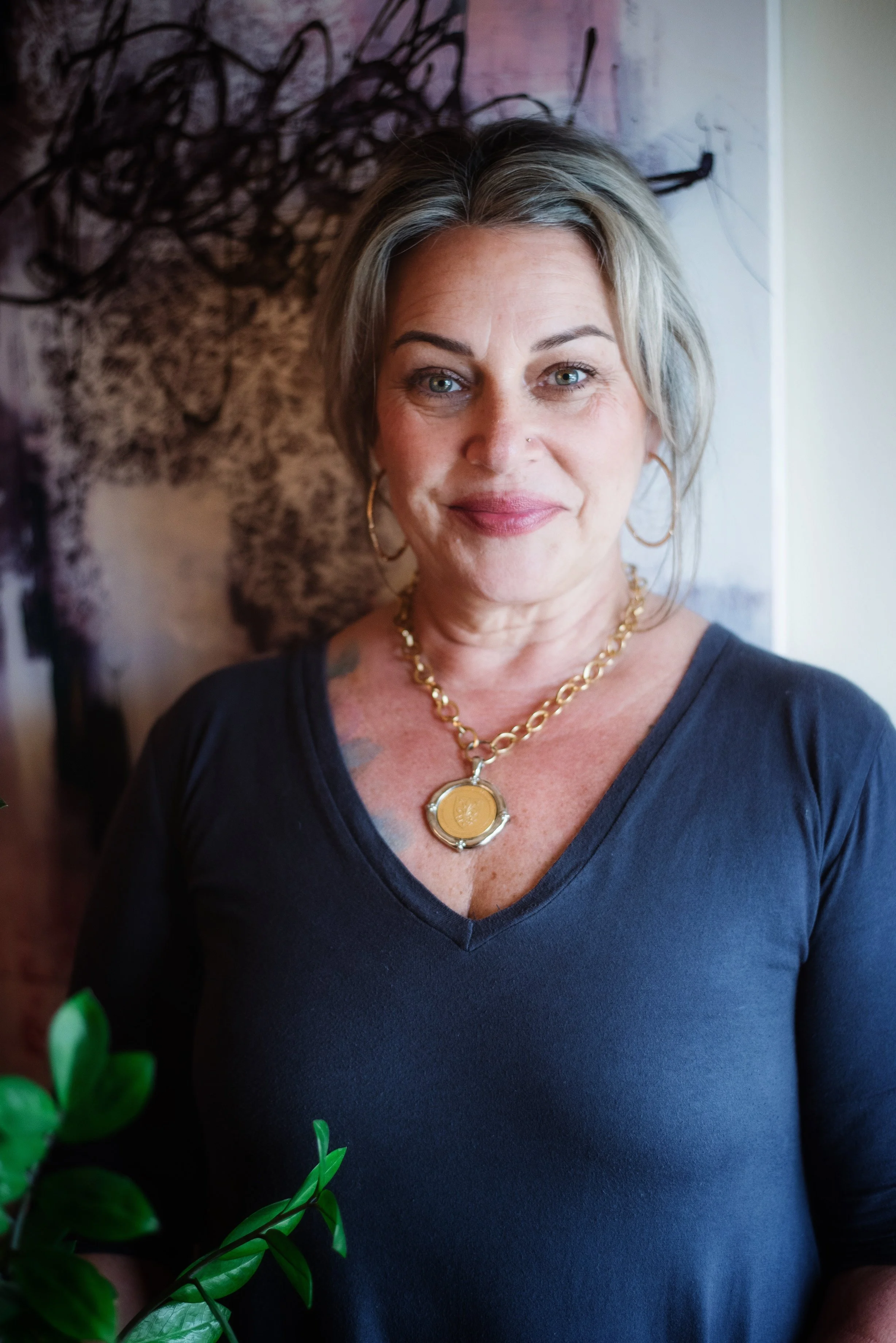 A woman with short gray hair, wearing large hoop earrings, a gold necklace with a circular pendant, and a black V-neck shirt, standing indoors with a colorful abstract background.