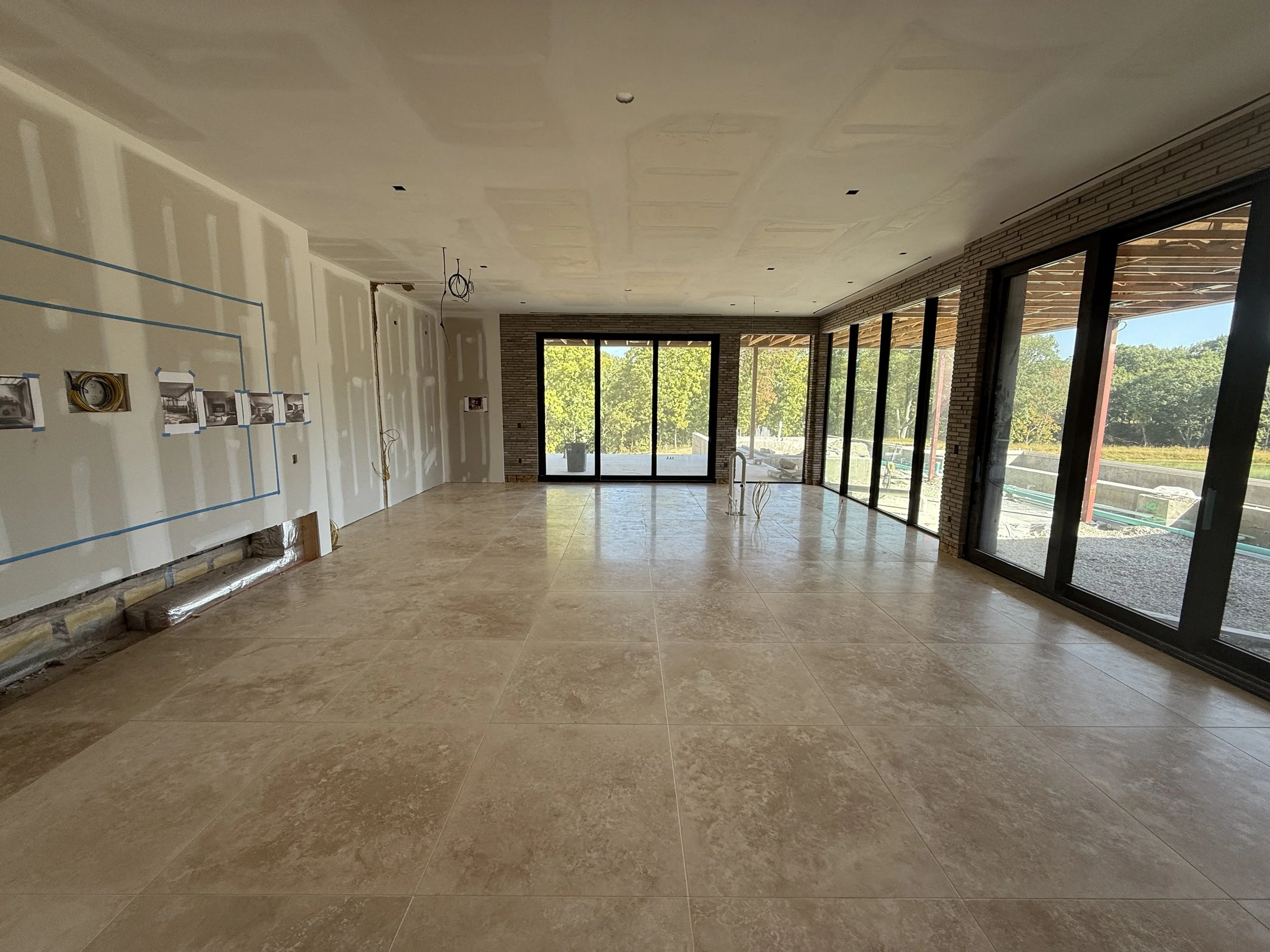 Empty room under construction with large beige tiled floor, unfinished walls with electrical wiring, and large floor-to-ceiling windows showing an outdoor view of trees and a construction area.