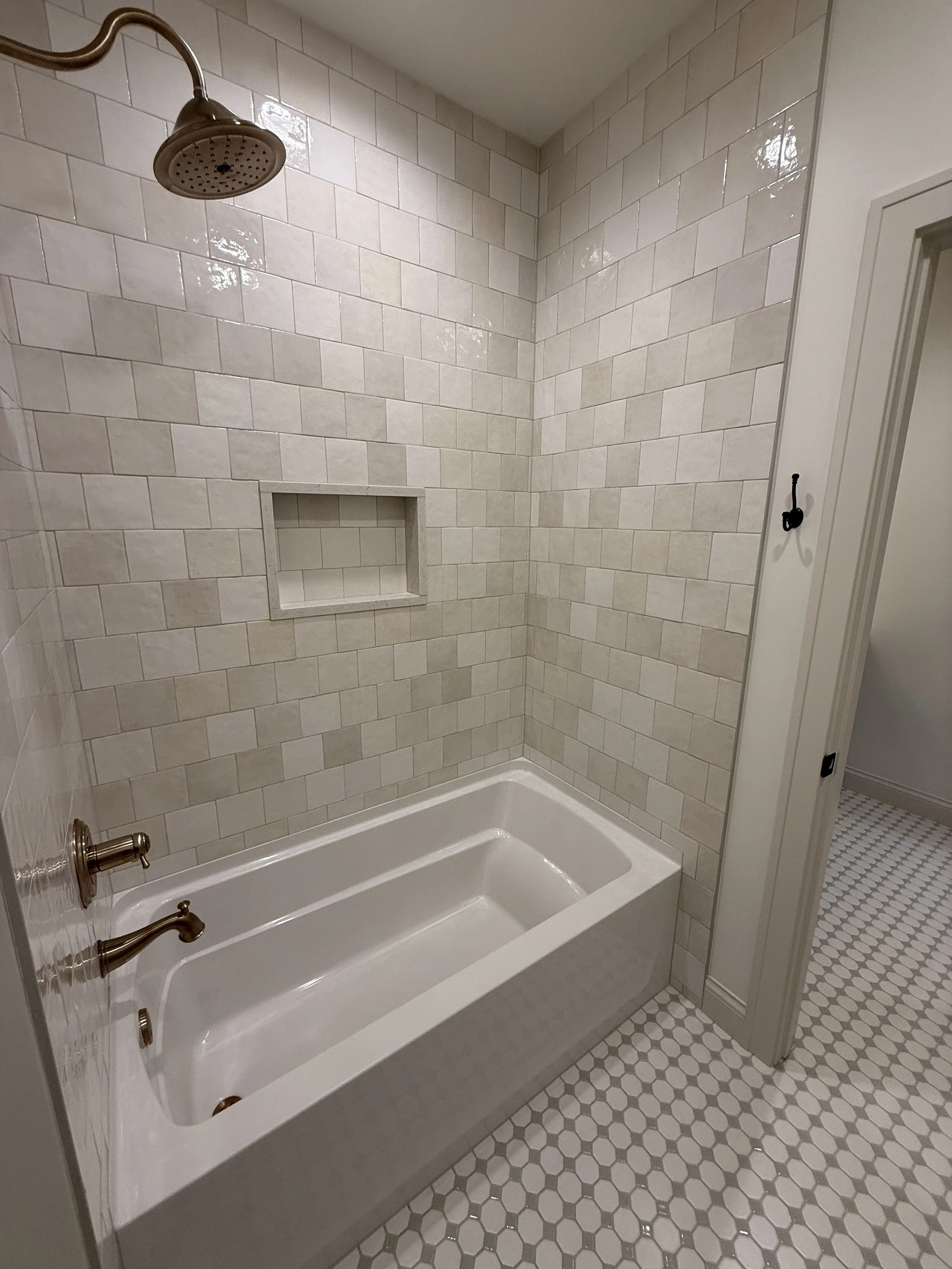 A bathroom shower area with beige square and rectangular tiles on the walls, a built-in niche, a bronze showerhead, and a bathtub with bronze fixtures. The floor has small hexagon tiles in white and light gray.
