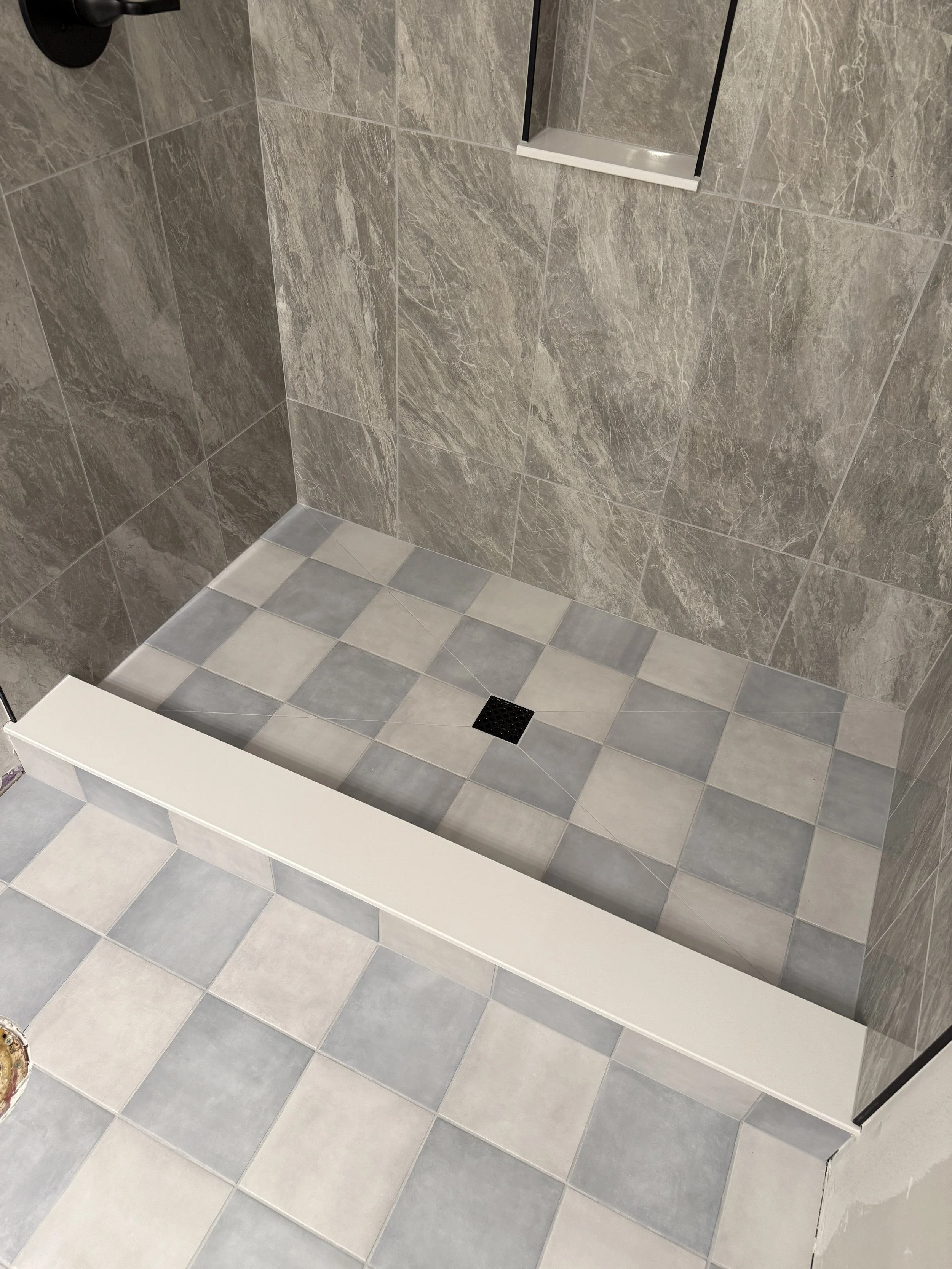 Shower with gray stone tile walls, gray and white checkered floor, a white shower curb, and a black drain on the floor.