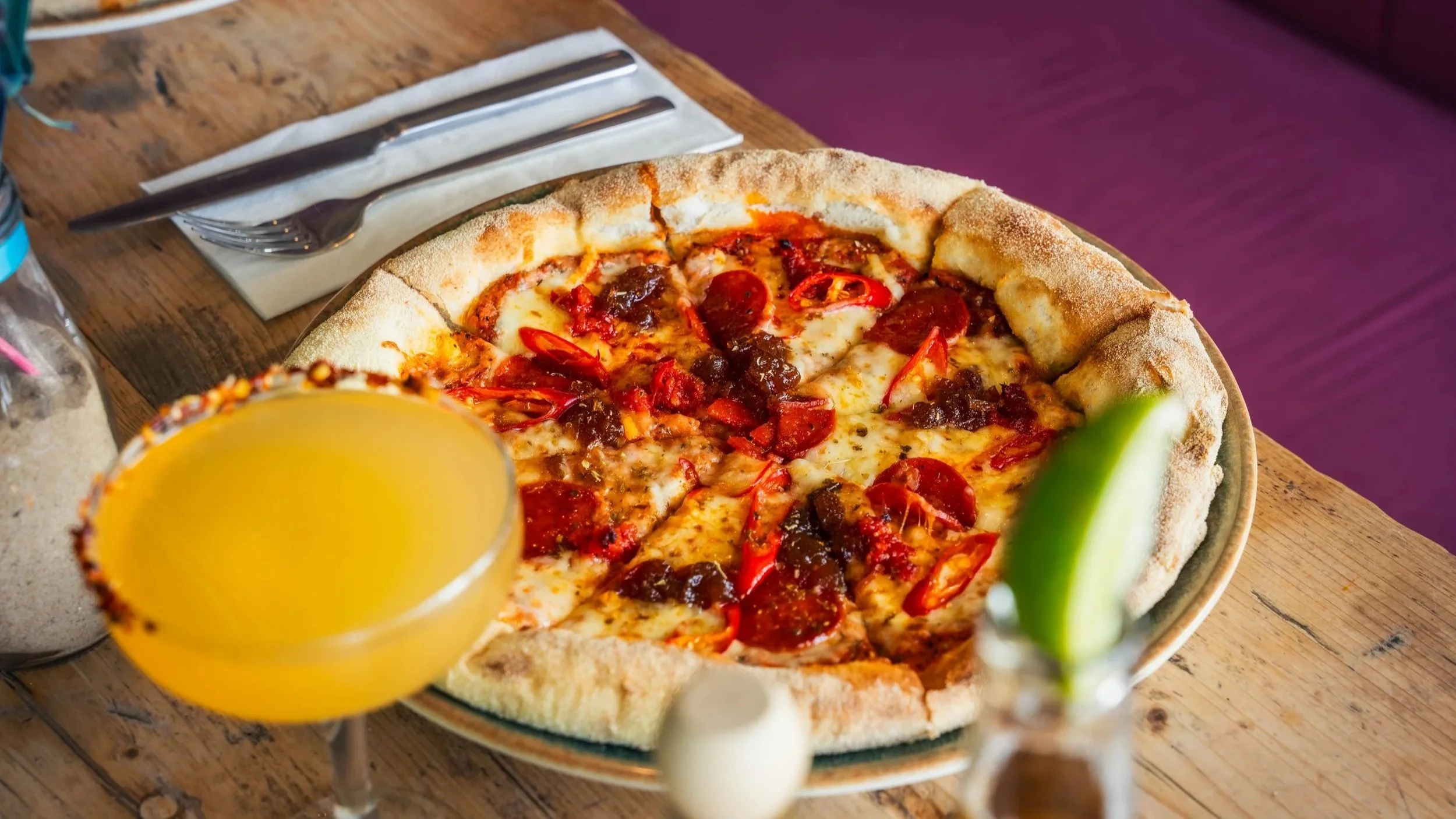 A pizza with pepperoni and red chili peppers on a wooden table, accompanied by a yellow cocktail with a salted rim and a lime wedge, and a small bottle of hot sauce.