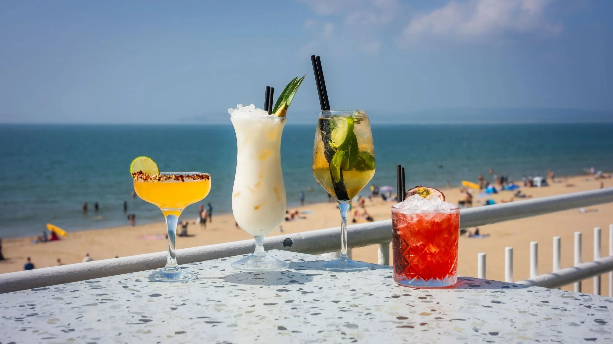 Four colorful cocktails on a table overlooking a beach with people, umbrellas, and the ocean under blue skies