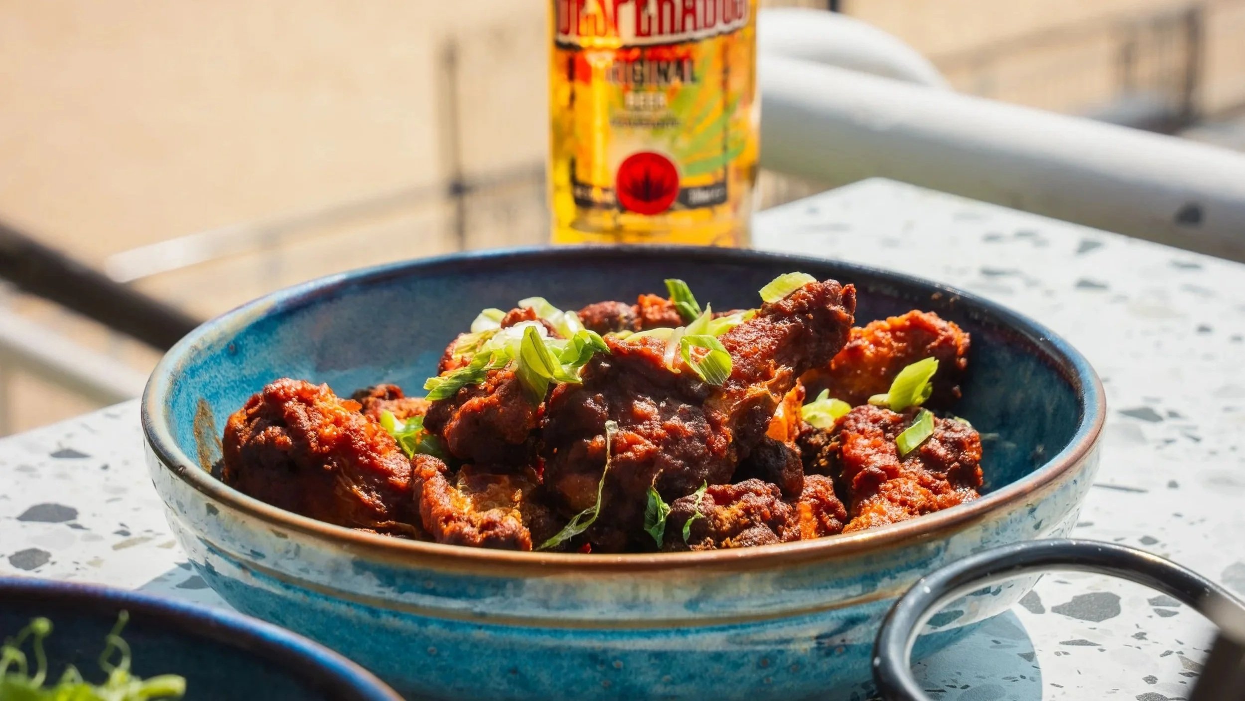 A blue bowl of spicy fried chicken garnished with chopped green onions on a white speckled marble table, with a can of juice or soda in the background.