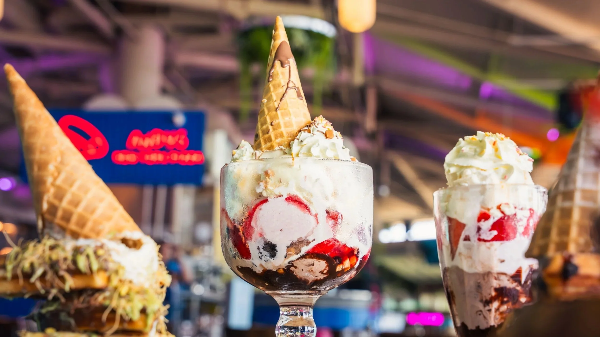 Close-up of a glass dessert cup filled with ice cream and topped with an ice cream cone, whipped cream, and sprinkles at a restaurant or cafe, with other ice cream dishes in the background.