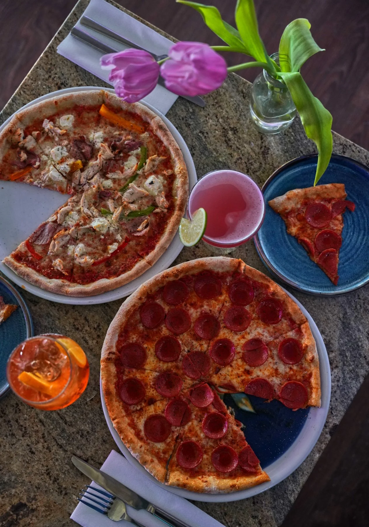 Two pizzas, one with pepperoni and one with chicken and vegetables, on a table with drinks and a tulip in a vase.