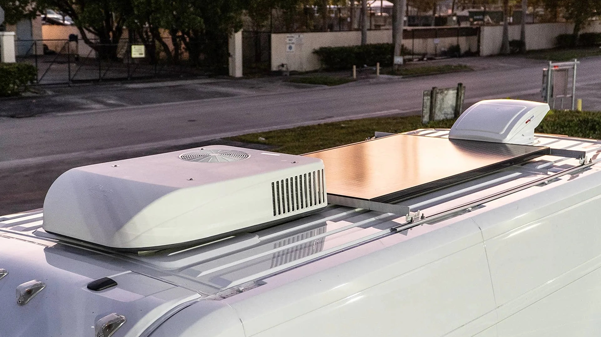 Close-up of the roof of a white vehicle, showing an rooftop air conditioning unit, a solar panel, and a roof vent in a parking lot.