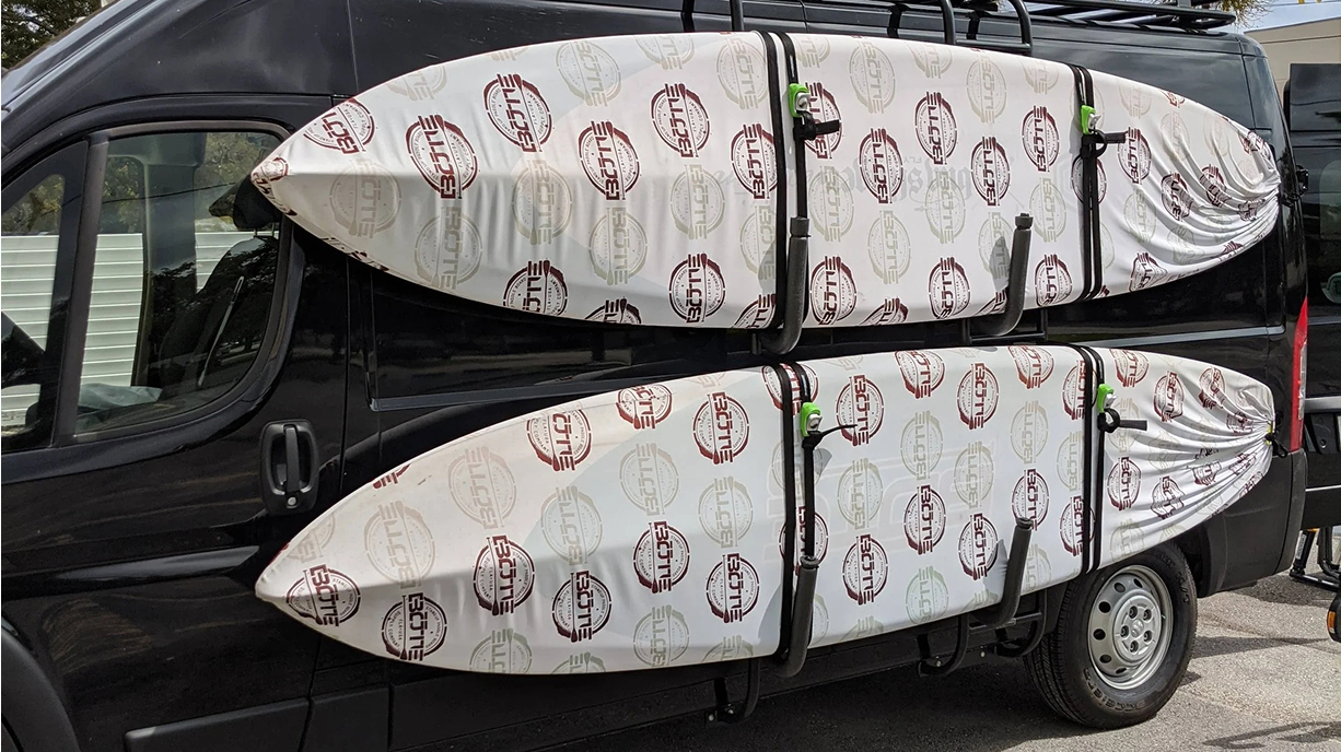 Black van with multiple white surfboards attached, each surfboard wrapped in protective covering with logos, secured to the side of the vehicle.