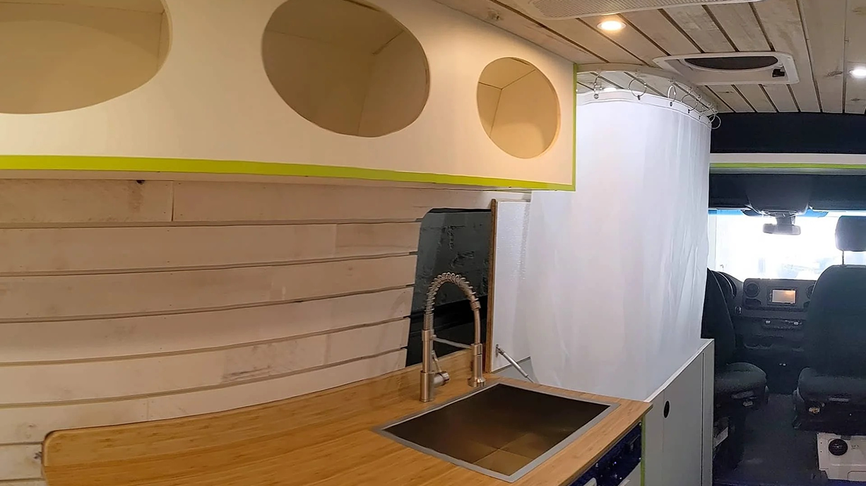 Interior of a tiny home or camper van with a wooden kitchen counter, sink, and a decorative wall with circular cutouts. The front seats and dashboard of the vehicle are visible in the background.