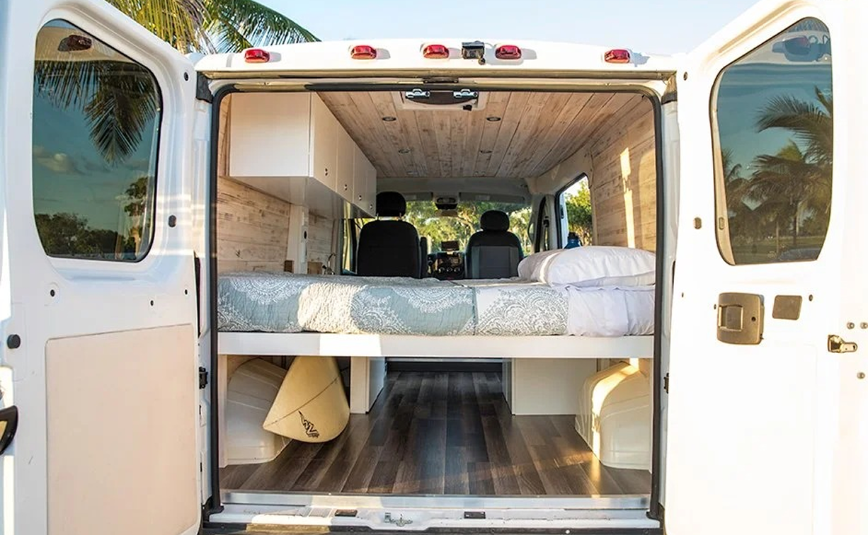 The interior of a white camper van with a bed and wooden ceiling and walls, and black driver and passenger seats visible from the back.