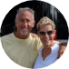 A man and a woman standing outdoors near a river, smiling at the camera with trees and water in the background.