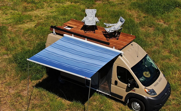 A beige camper van with a blue and white striped awning parked on grass. The RV has a wooden deck on top with two chairs and a small table.