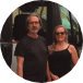 Two people standing outdoors, posing for a photo. The person on the left is wearing a black shirt and sunglasses, and the person on the right is wearing glasses and a black dress.