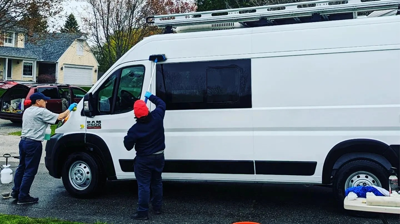 Two men cleaning a white van outdoors in a suburban neighborhood.