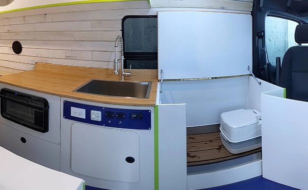 Interior of a camper van with a small kitchen area and a fold-up bed with a portable toilet.