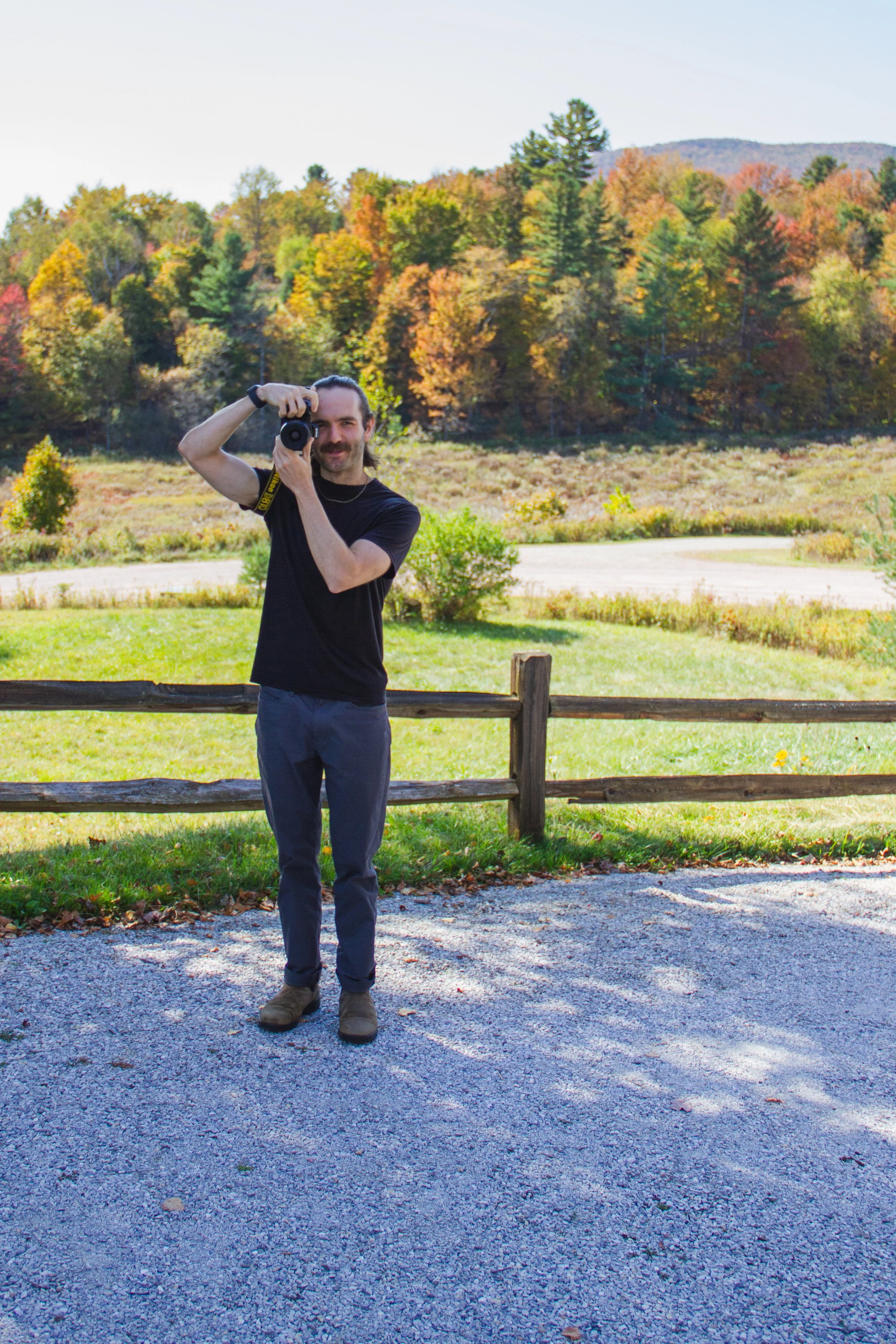 A man with dark hair and a mustache, wearing a black t-shirt, navy pants, and boots, holding a camera near his face, standing outdoors in front of a wooden fence with a background of colorful fall foliage and trees.