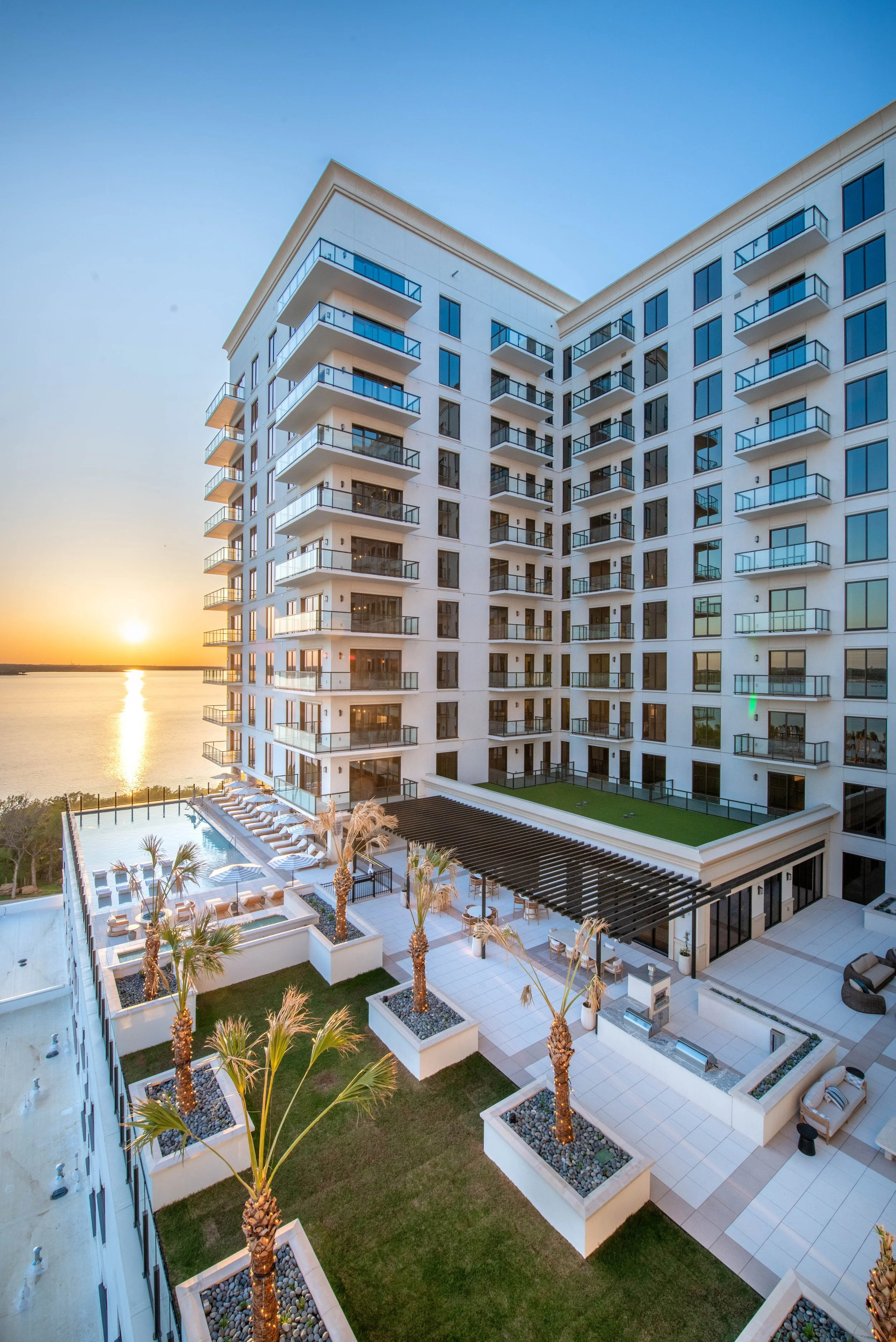 Modern high-rise apartment building with outdoor pool area, palm trees, and sunset view over water.