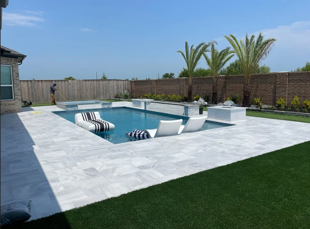 Backyard with a swimming pool surrounded by white marble tiles, with two modern white lounge chairs and a striped pool float. There are three palm trees along the brick wall, which has landscaping with plants and rocks. A person is cleaning the yard in the background.