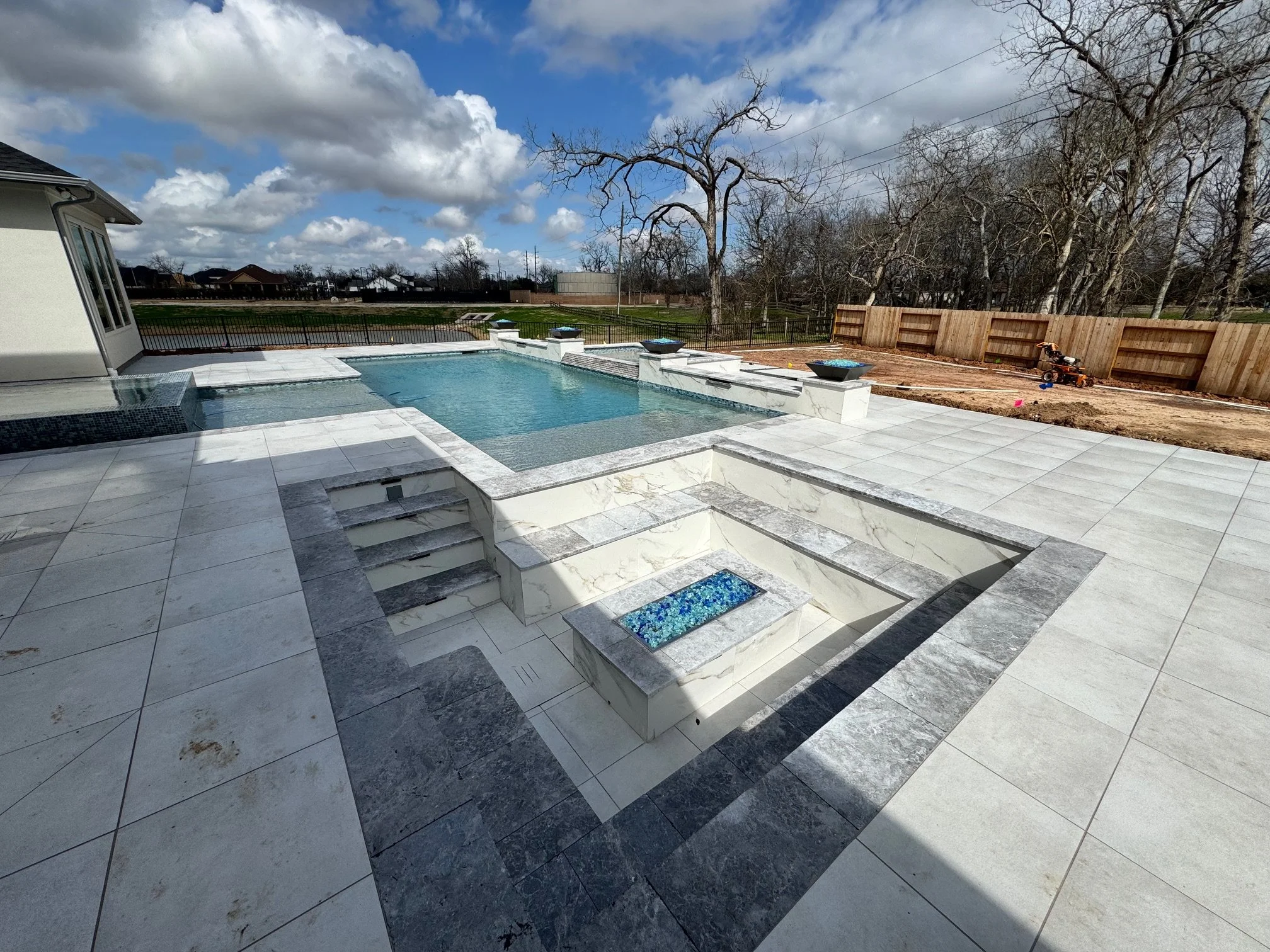 New backyard with a swimming pool, spa, and tiled patio, enclosed by a fence, with trees in the background under partly cloudy sky.