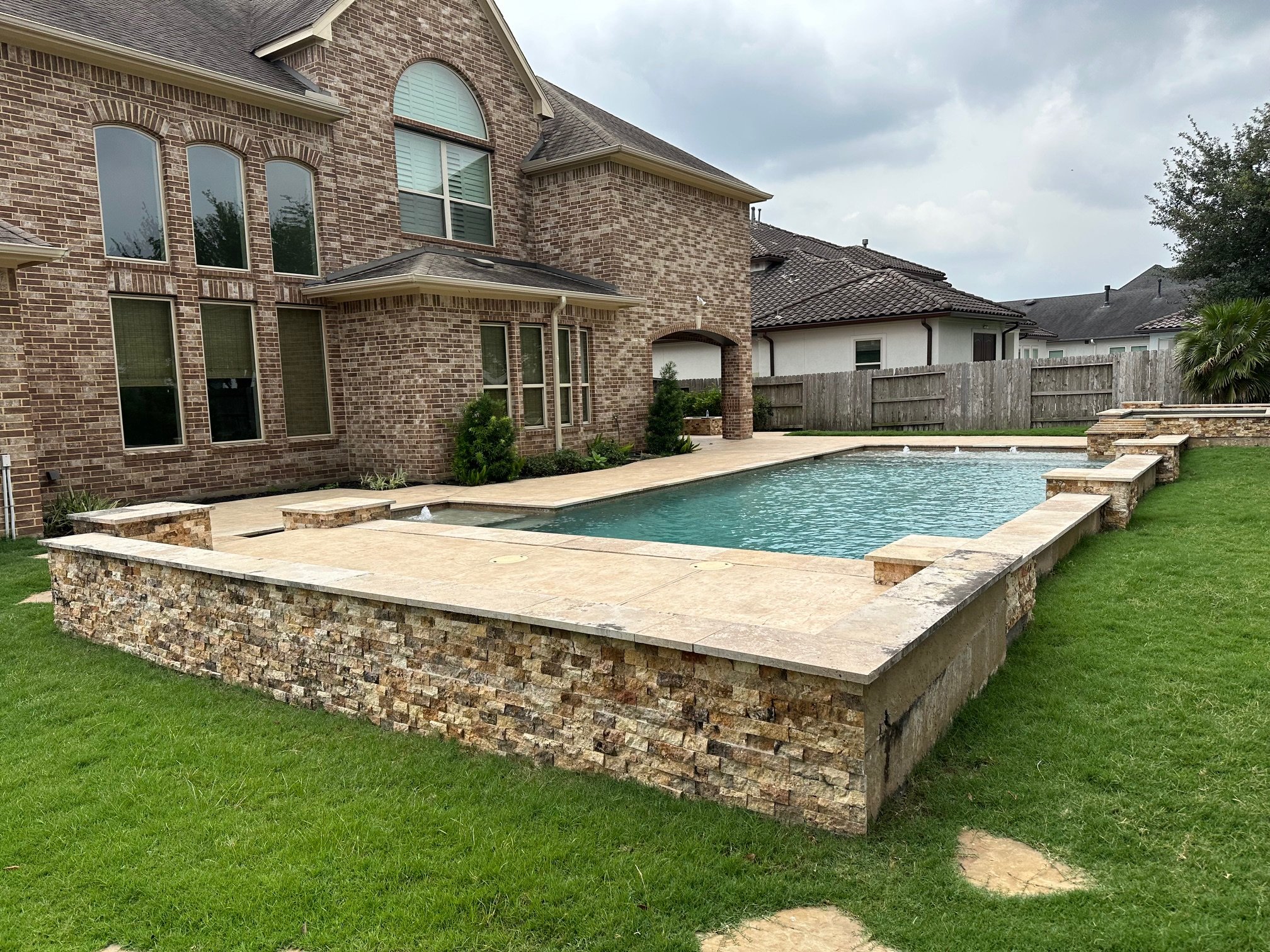 Backyard with a swimming pool surrounded by a stone deck, adjacent to a brick house with multiple windows, and a wooden fence in the background under a cloudy sky.