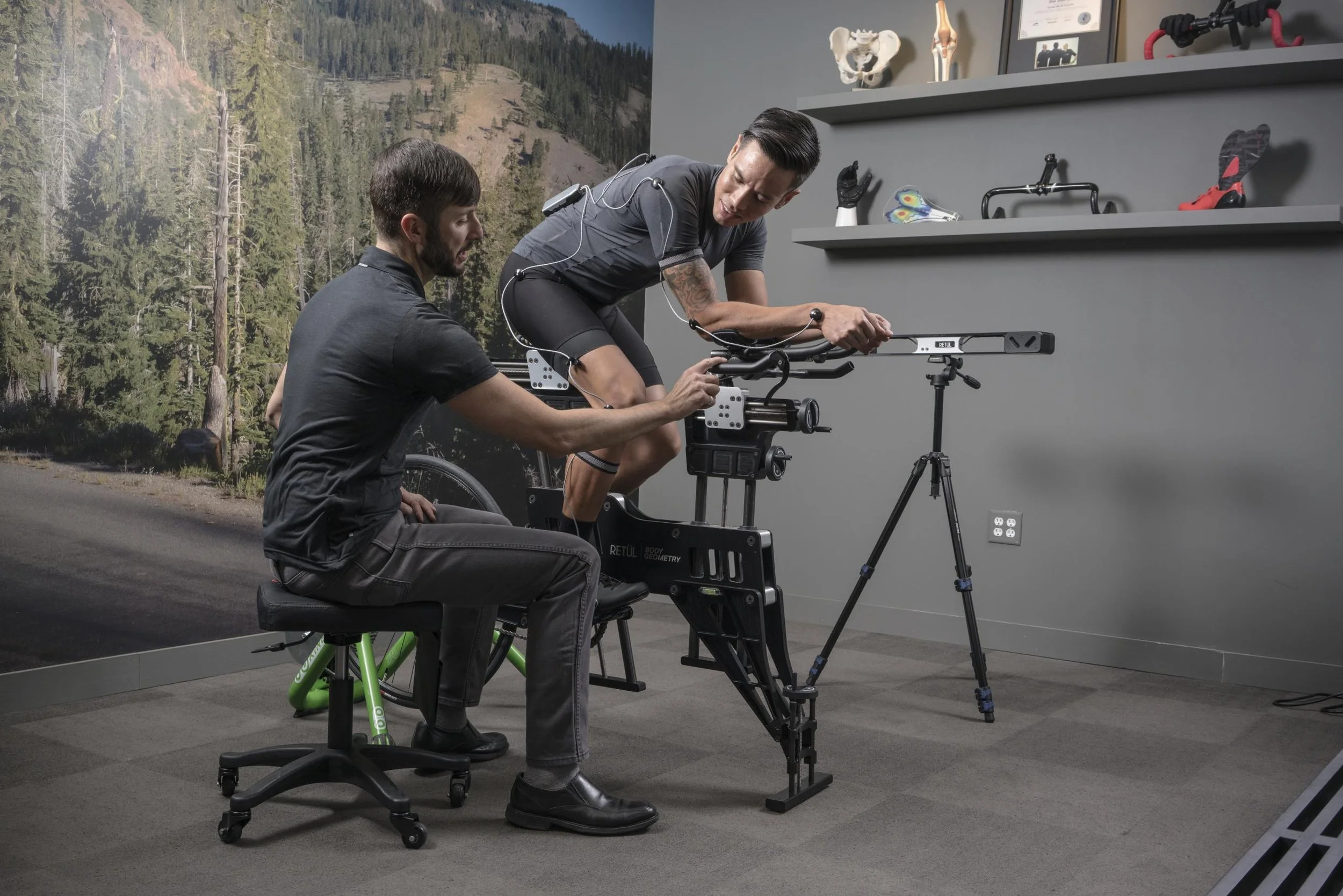 A person is being fitted on a bicycle fitting system by a technician in a room with cycling equipment and a nature backdrop. The cyclist is wearing sensors and athletic gear.