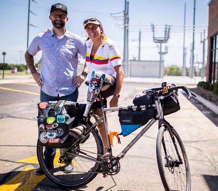 Two people standing outdoors with a bicycle, smiling. One person is wearing a cycling jersey and the other is in casual clothes. The bicycle is equipped with bags and decorated with various stickers. They are in an urban setting with buildings and power lines visible in the background.