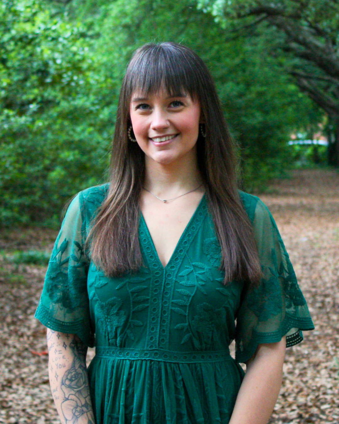 Smiling woman with long hair and bangs, wearing a floral patterned blouse, standing outdoors with greenery in the background.