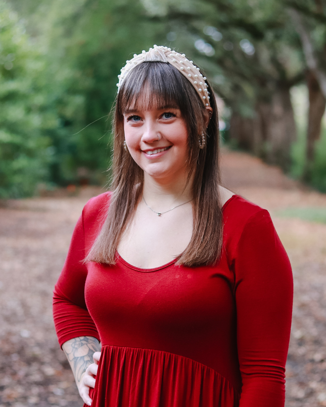 Smiling woman with long hair and bangs, wearing a floral patterned blouse, standing outdoors with greenery in the background.