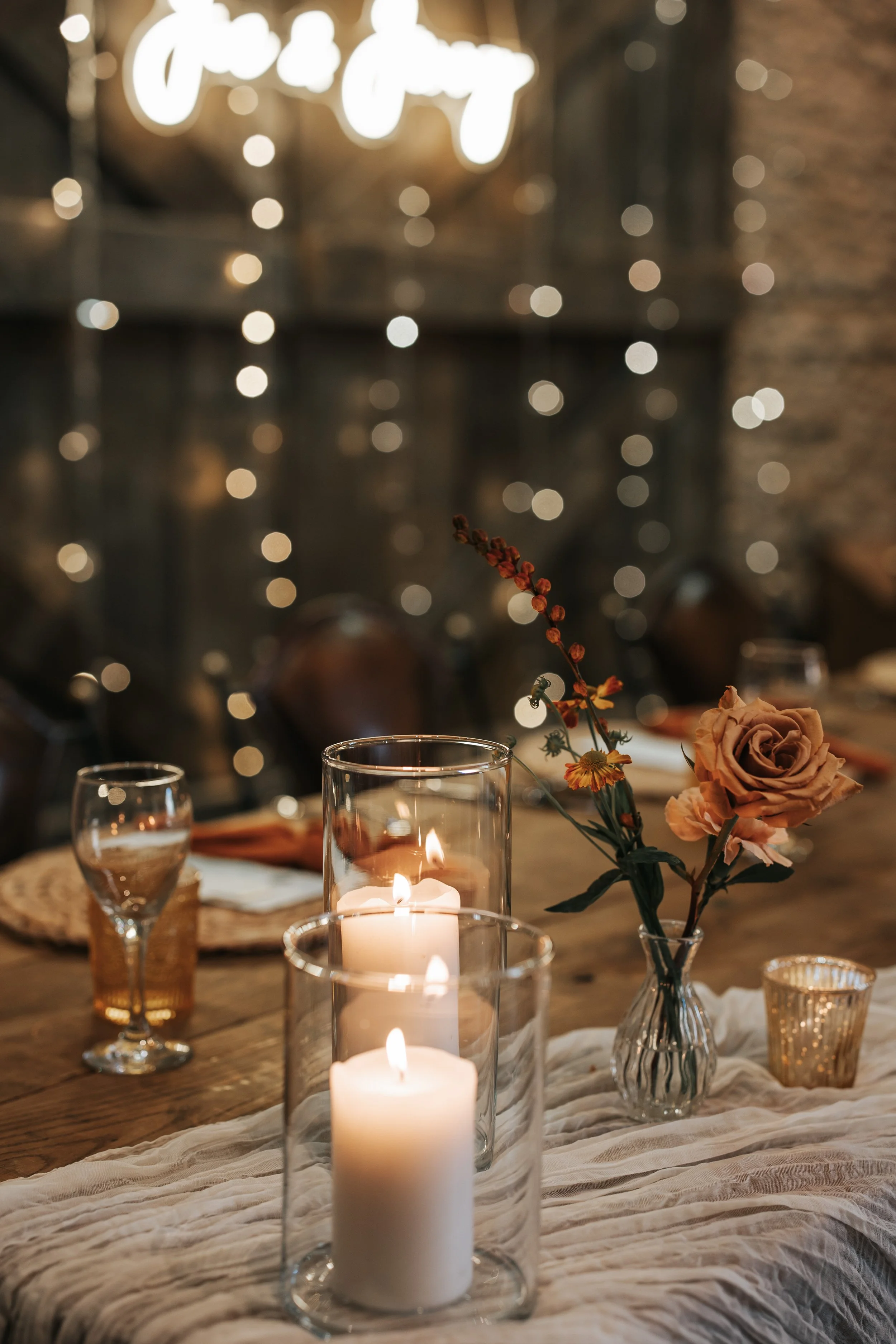 A warmly lit dining table decorated with lit candles in glass holders, a small vase with a beige and rust-colored flower and greenery, and gold-colored glasses, set against a background with bokeh lights and a illuminated sign.