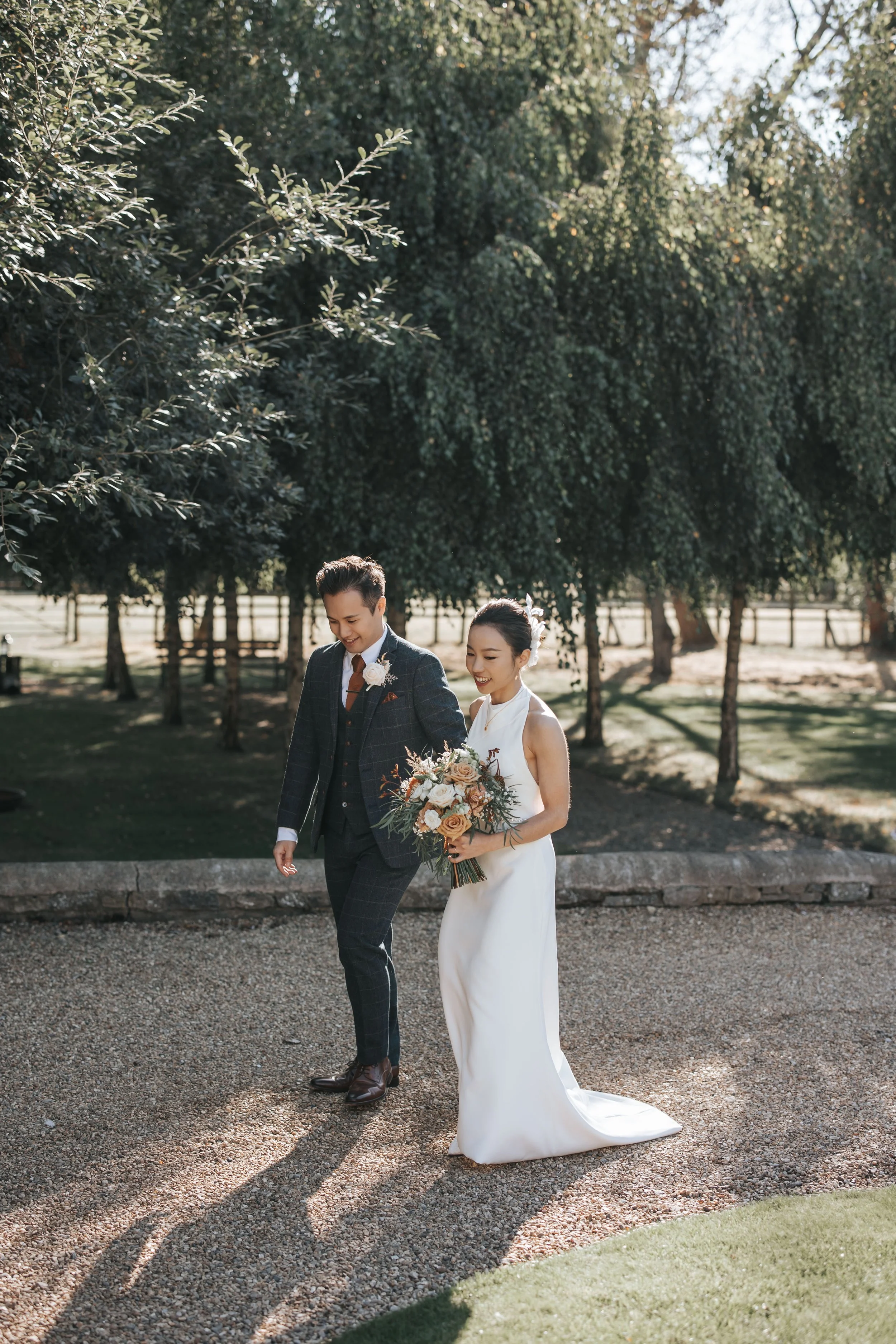 A bride and groom walking outdoors on a gravel path surrounded by trees during their wedding, with the bride holding a bouquet of flowers.