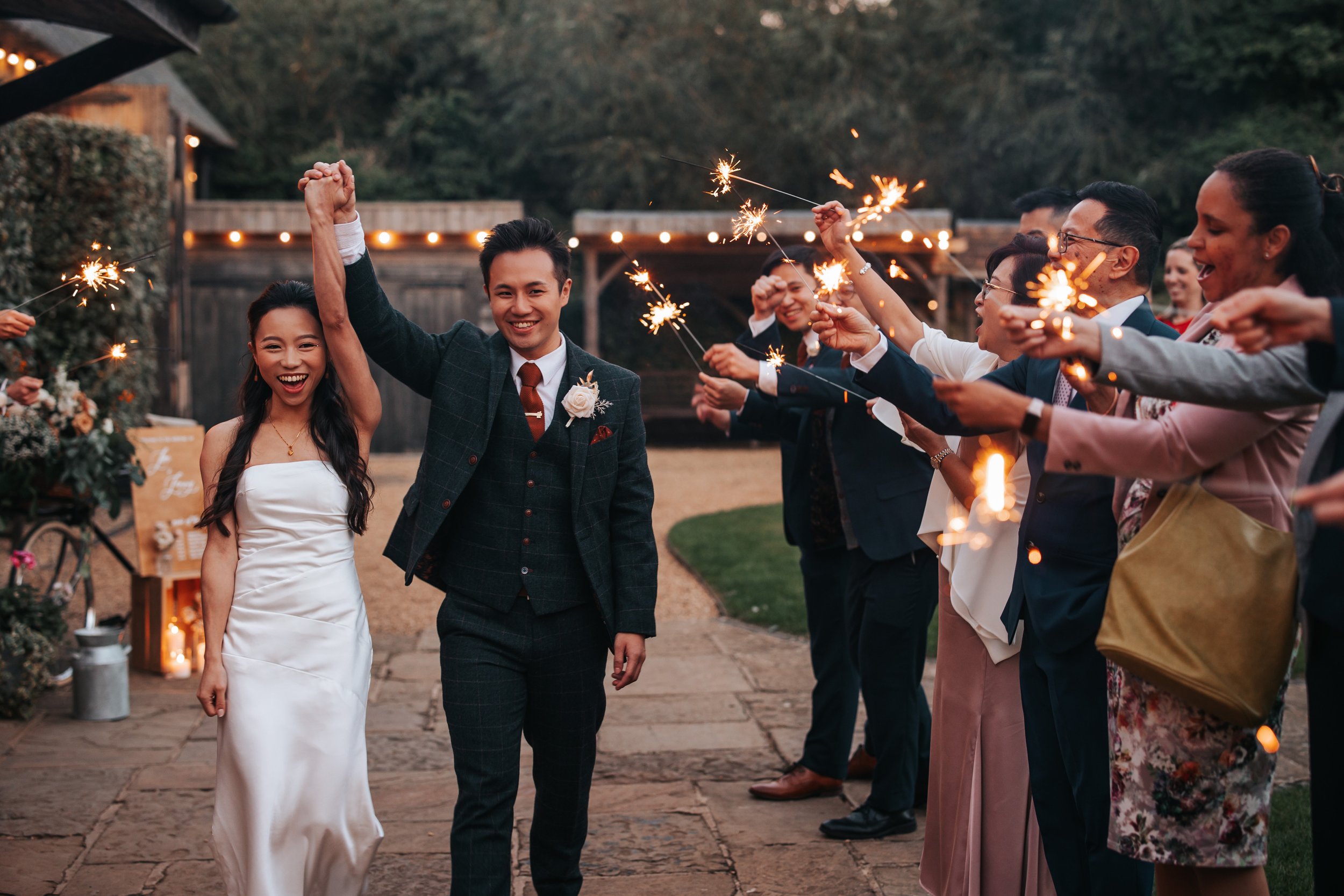 Couple celebrating wedding with sparklers, bride in white gown, groom in suit, surrounded by friends and family at outdoor evening event.
