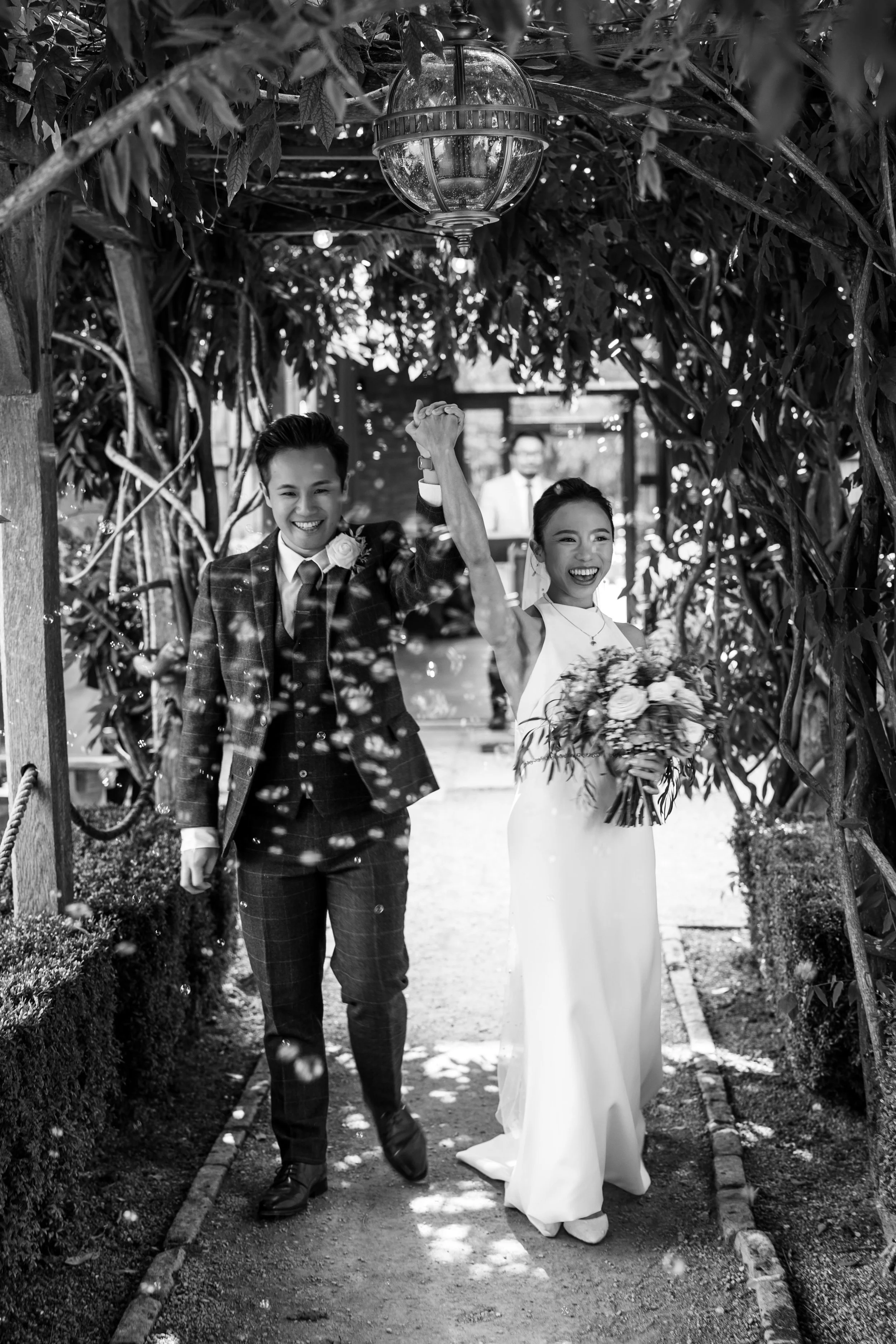Black and white photo of a newly married couple celebrating their wedding outdoors. The bride and groom are holding hands and smiling as they walk through a garden archway, with the bride holding a bouquet of flowers.