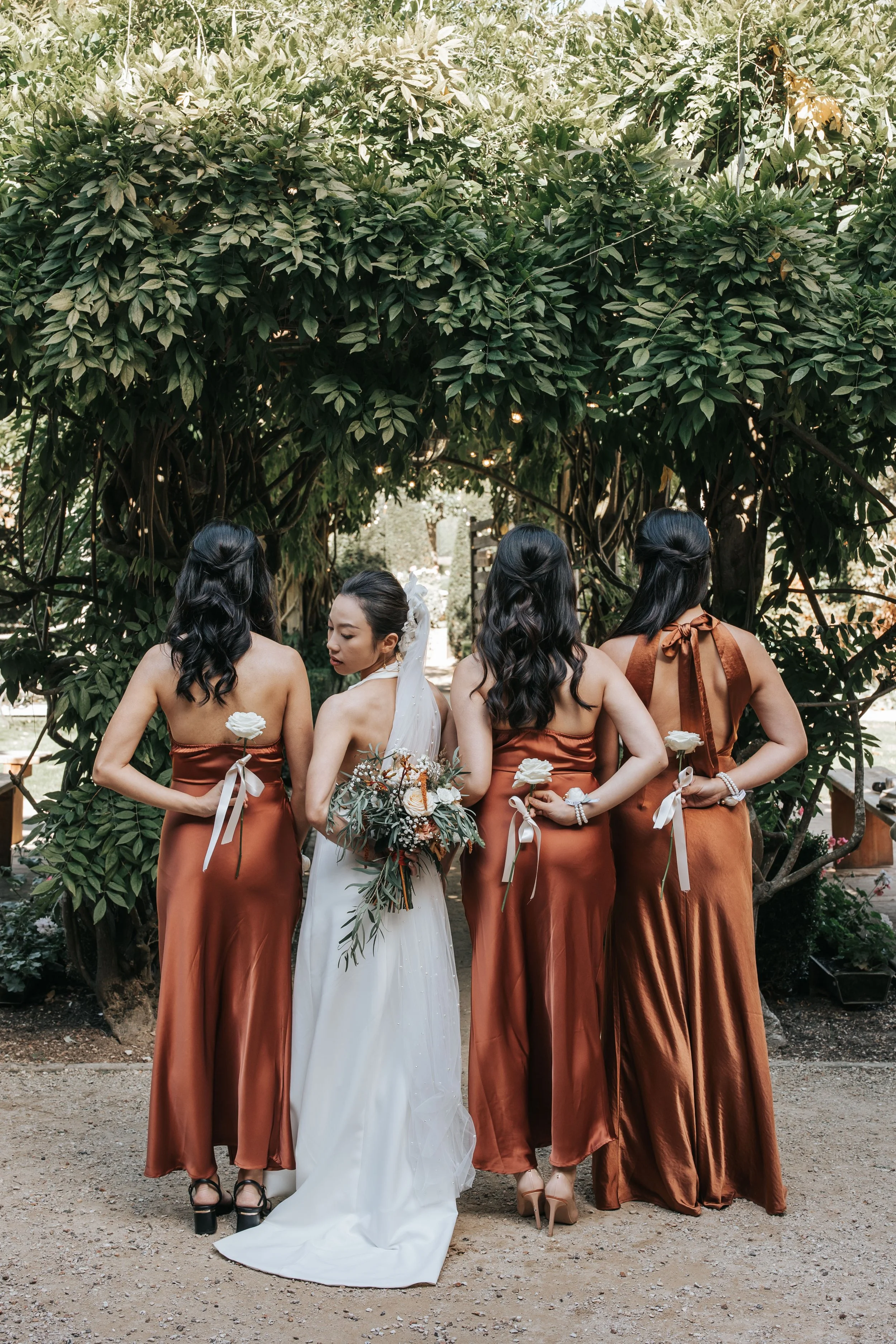 A bride and three bridesmaids posing outdoors in front of a large leafy tree. The bridesmaids are wearing matching brown satin dresses and holding white flowers.
