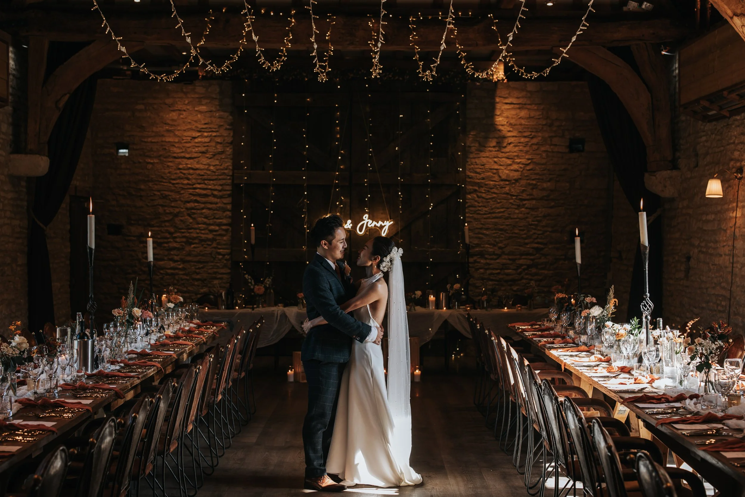 A bride and groom dancing in a warmly lit rustic wedding reception hall decorated with string lights, candles, and floral centerpieces on long banquet tables.