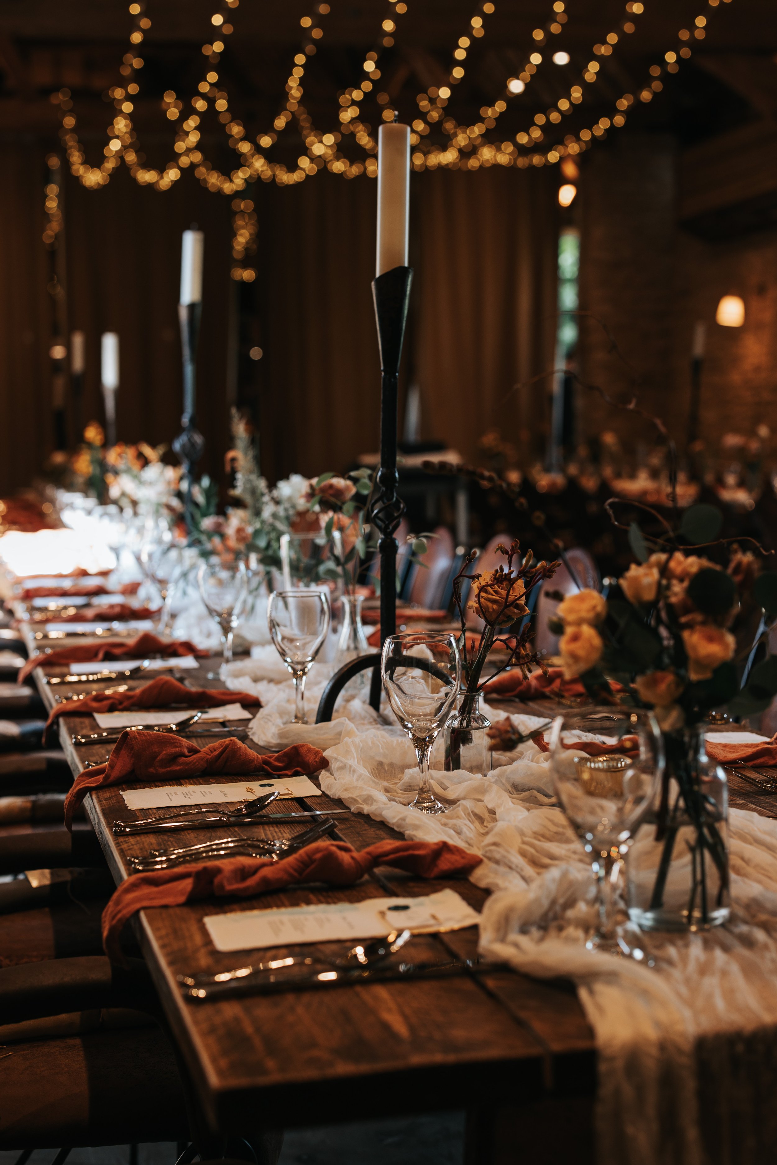 Elegant indoor wedding reception table decorated with candles, flowers, glassware, and rustic table runner.