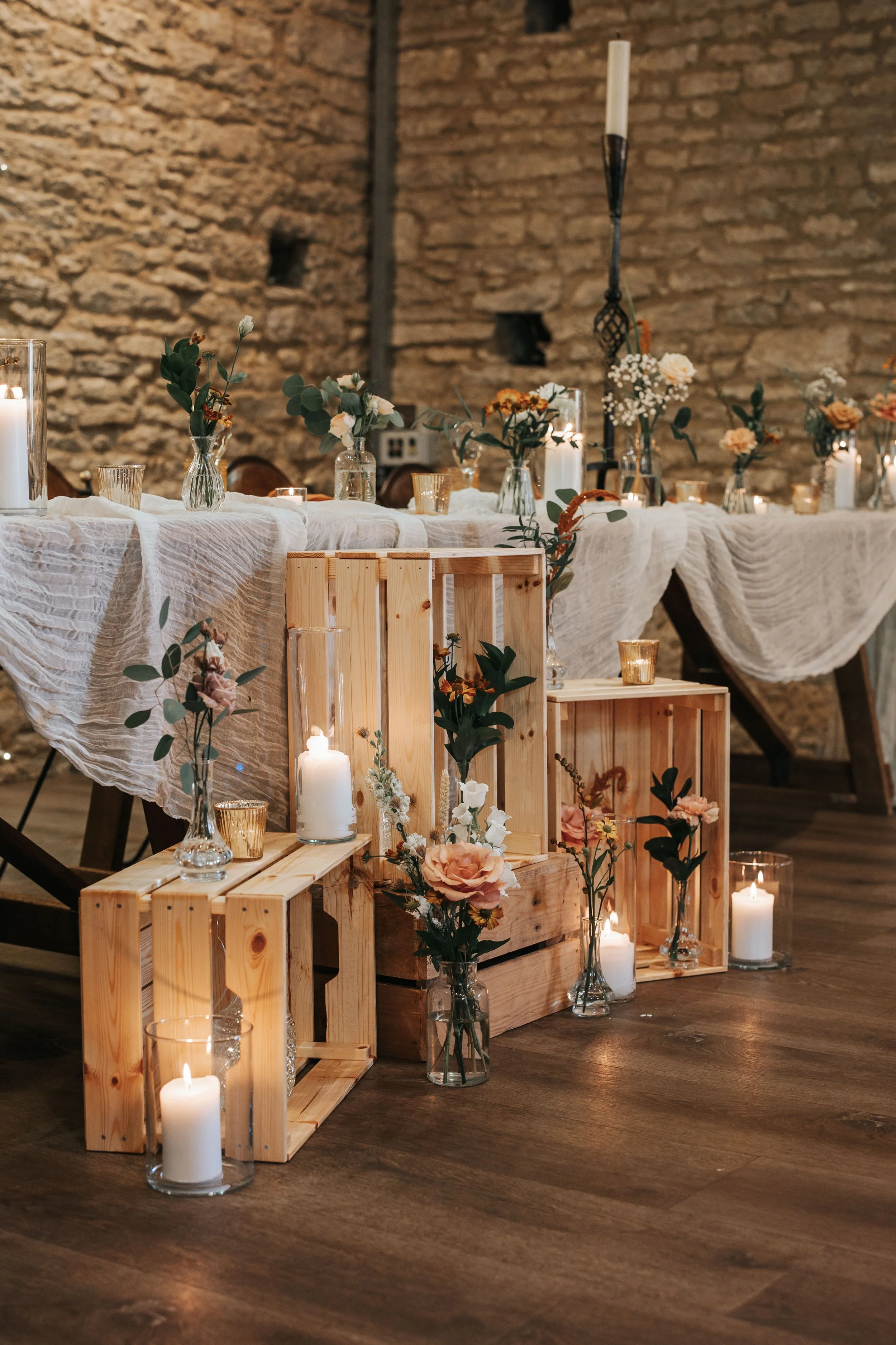 Decorated table with candles and flowers, rustic wooden crates, and a beige tablecloth in a room with brick walls.