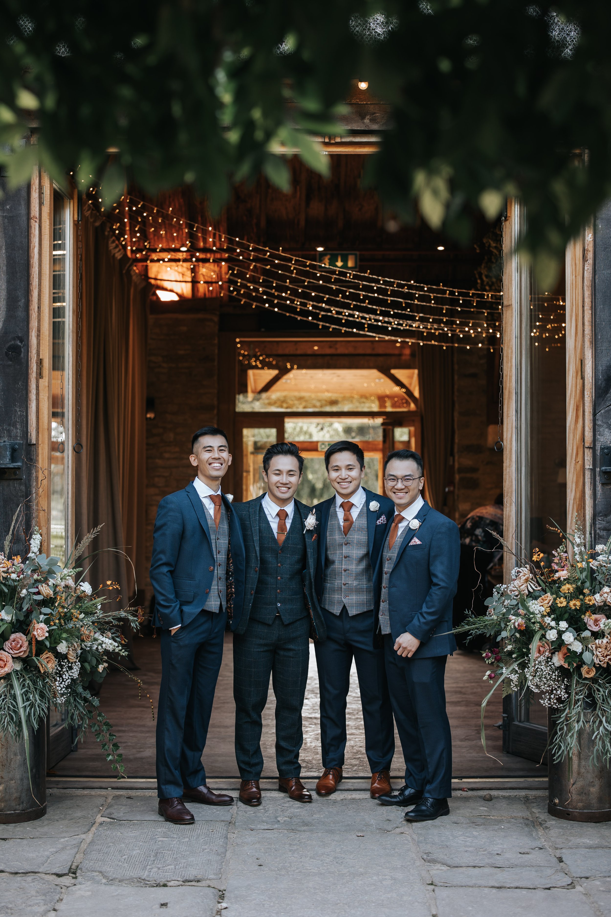 Four men dressed in suits standing together at a wedding or formal event, smiling at the camera, with decorated doorway and fairy lights above them.