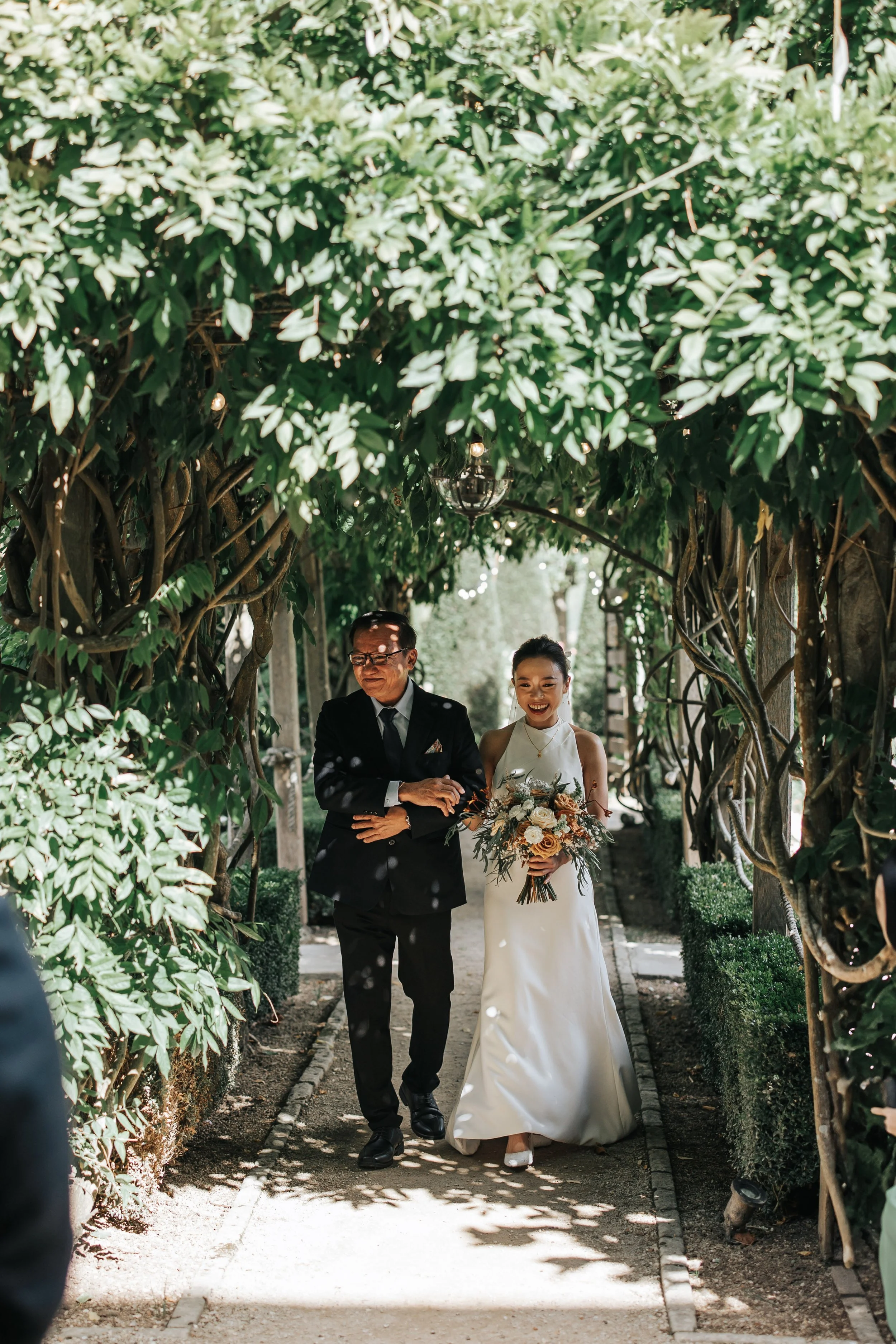 A bride walking down a garden aisle arm-in-arm with her father, holding a bouquet of flowers, smiling.
