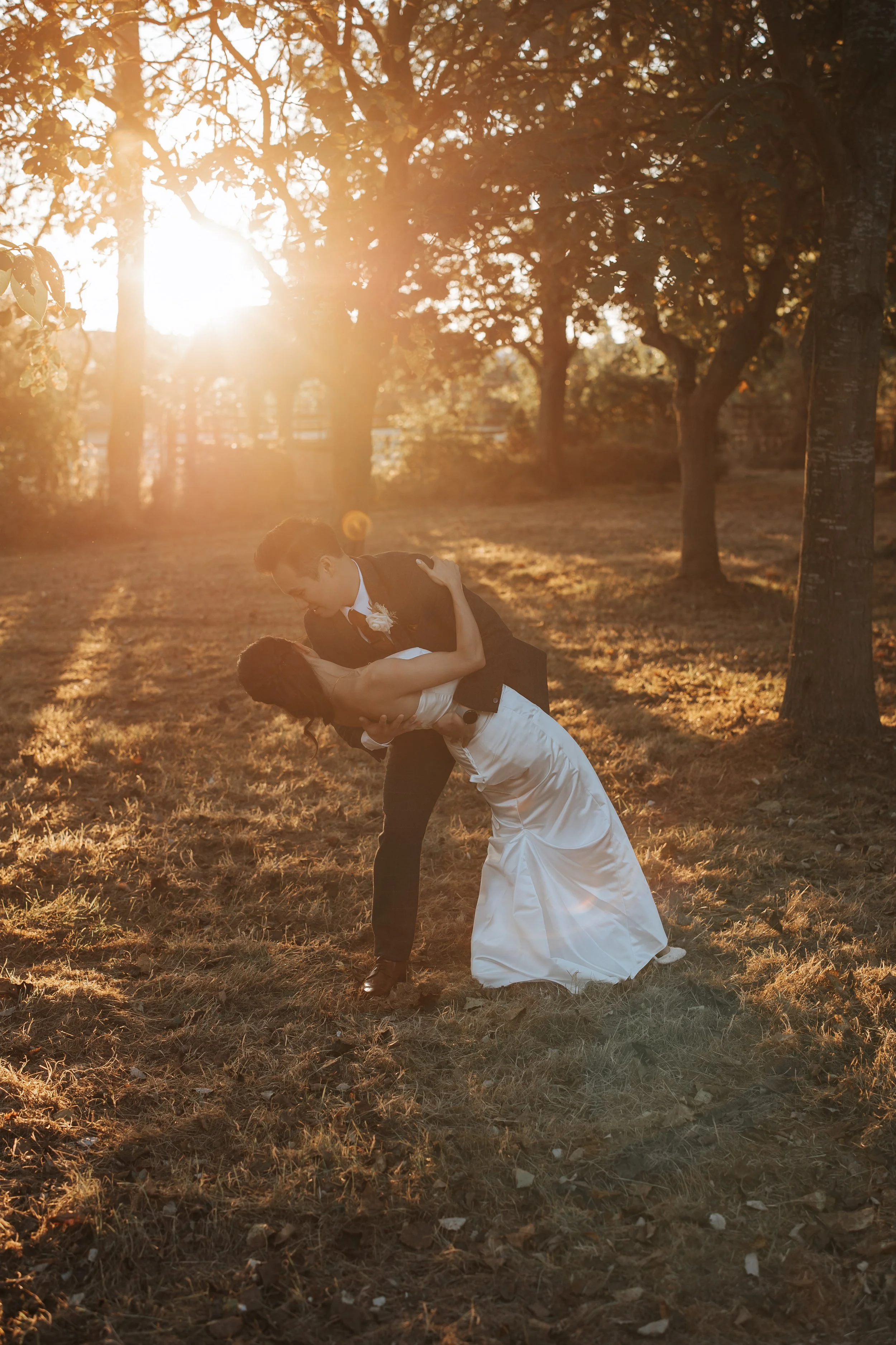 A couple in wedding attire dancing outdoors at sunset, surrounded by trees and golden light.