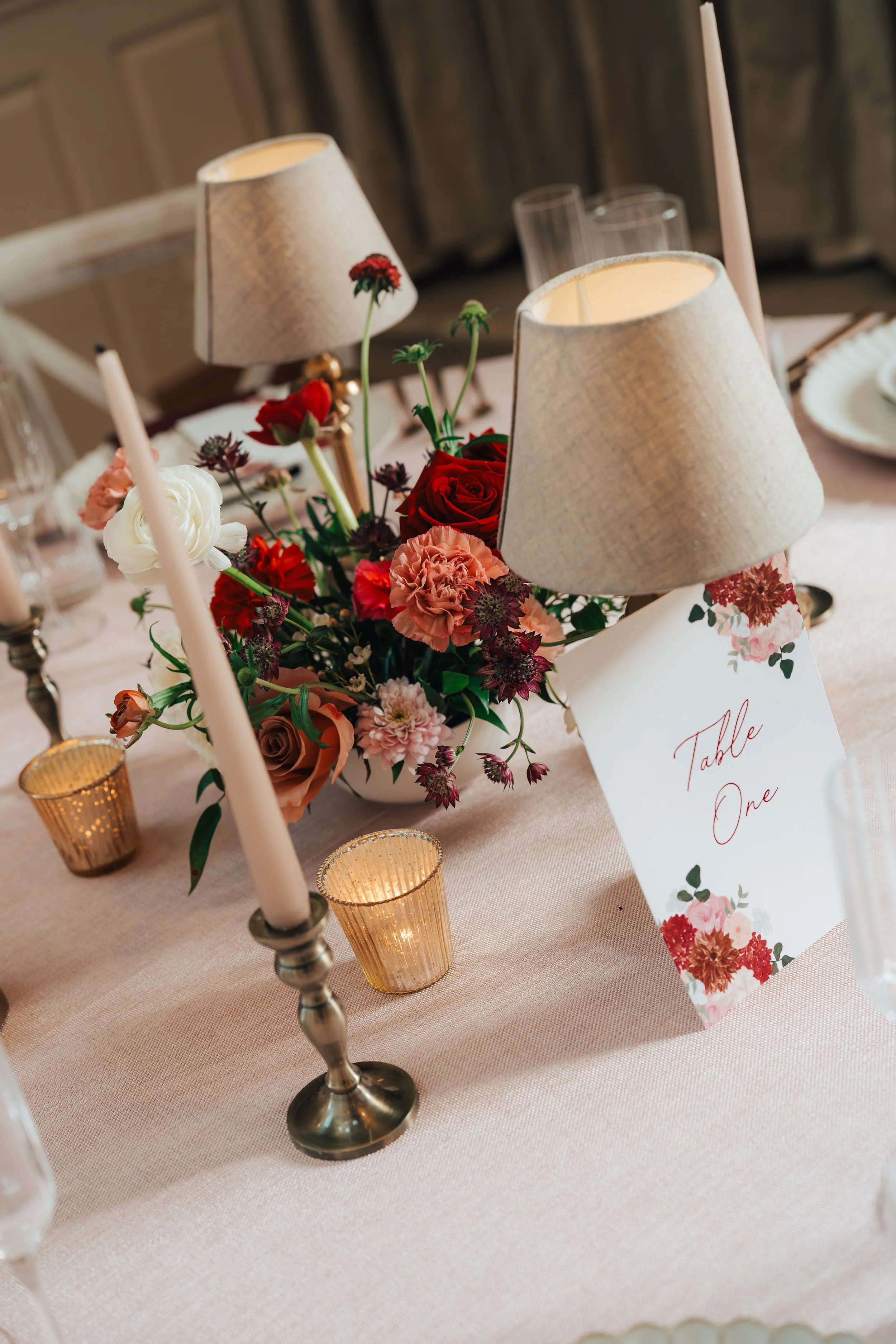 Red, peach and white wedding table flowers arranged alongside candles and small table lamps.