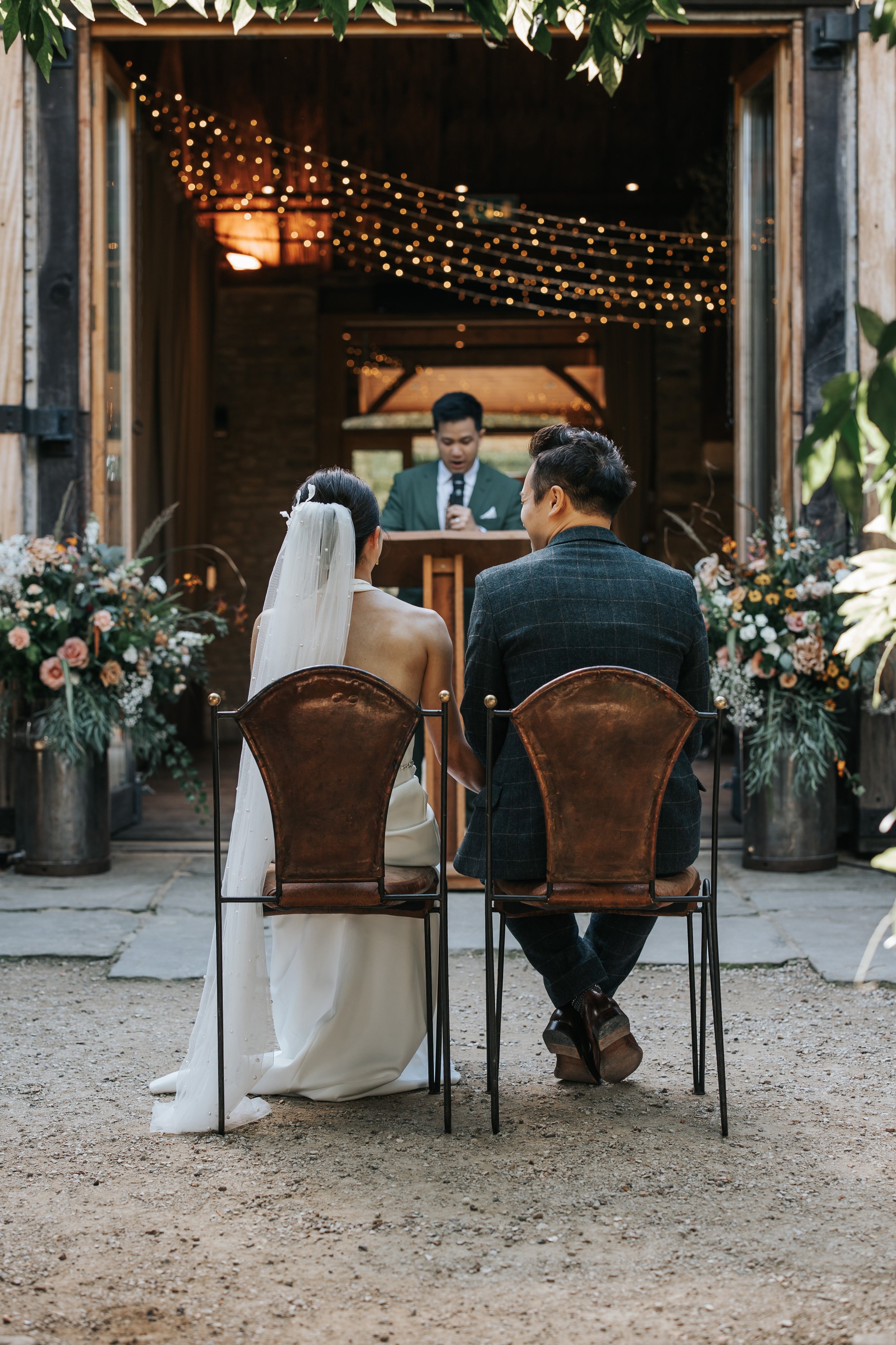 A bride and groom sitting side by side during their wedding ceremony outdoors, facing an officiant. The bride wears a white wedding dress and veil, and the groom wears a dark checked suit. There are floral arrangements on both sides and string lights