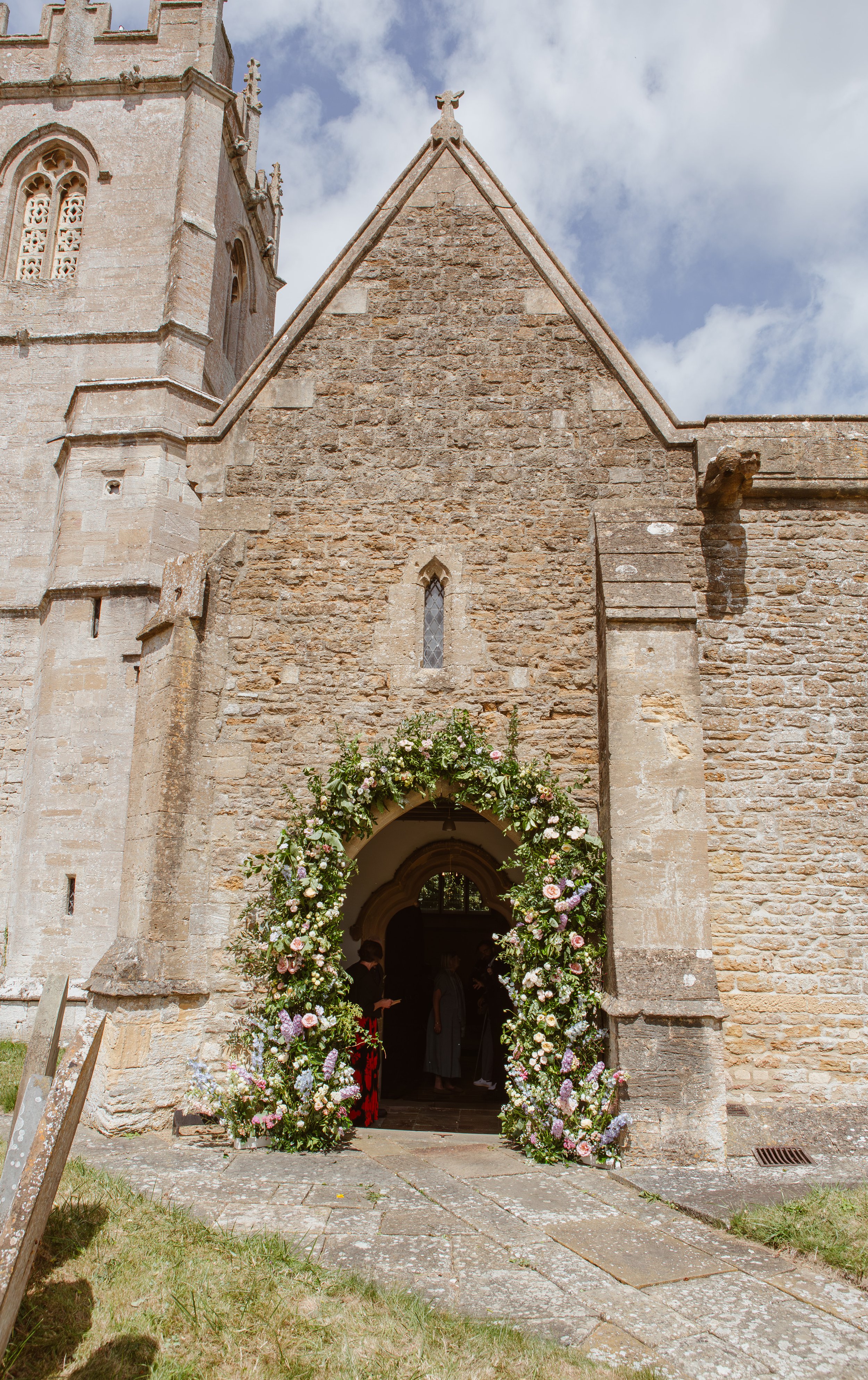 Pink and lilac wedding flower arch with greenery framing a stone church entrance.